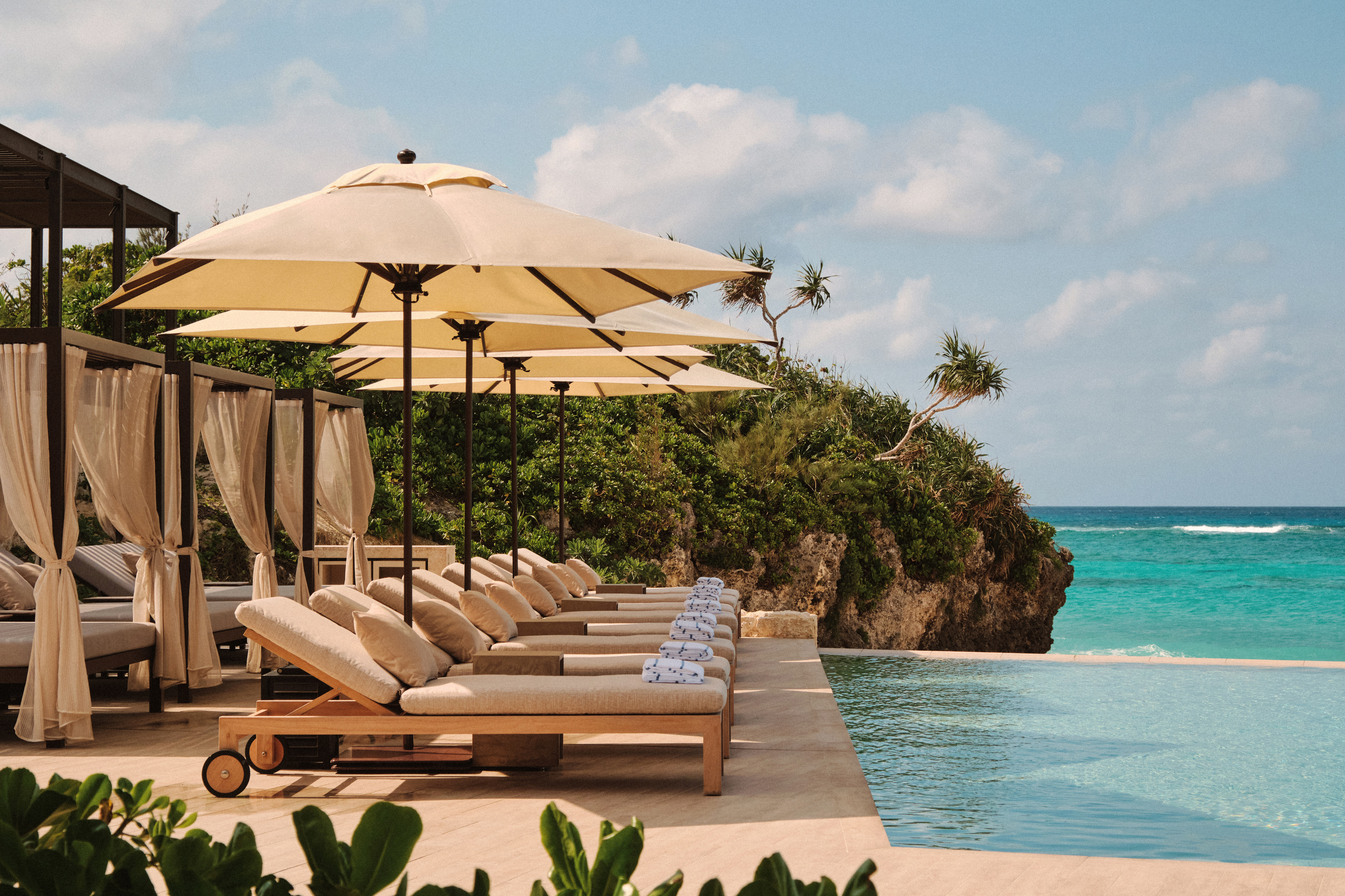 Cream loungers and umbrellas line an infinity pool at Rosewood Miyakojima, overlooking bright blue coastal water.
