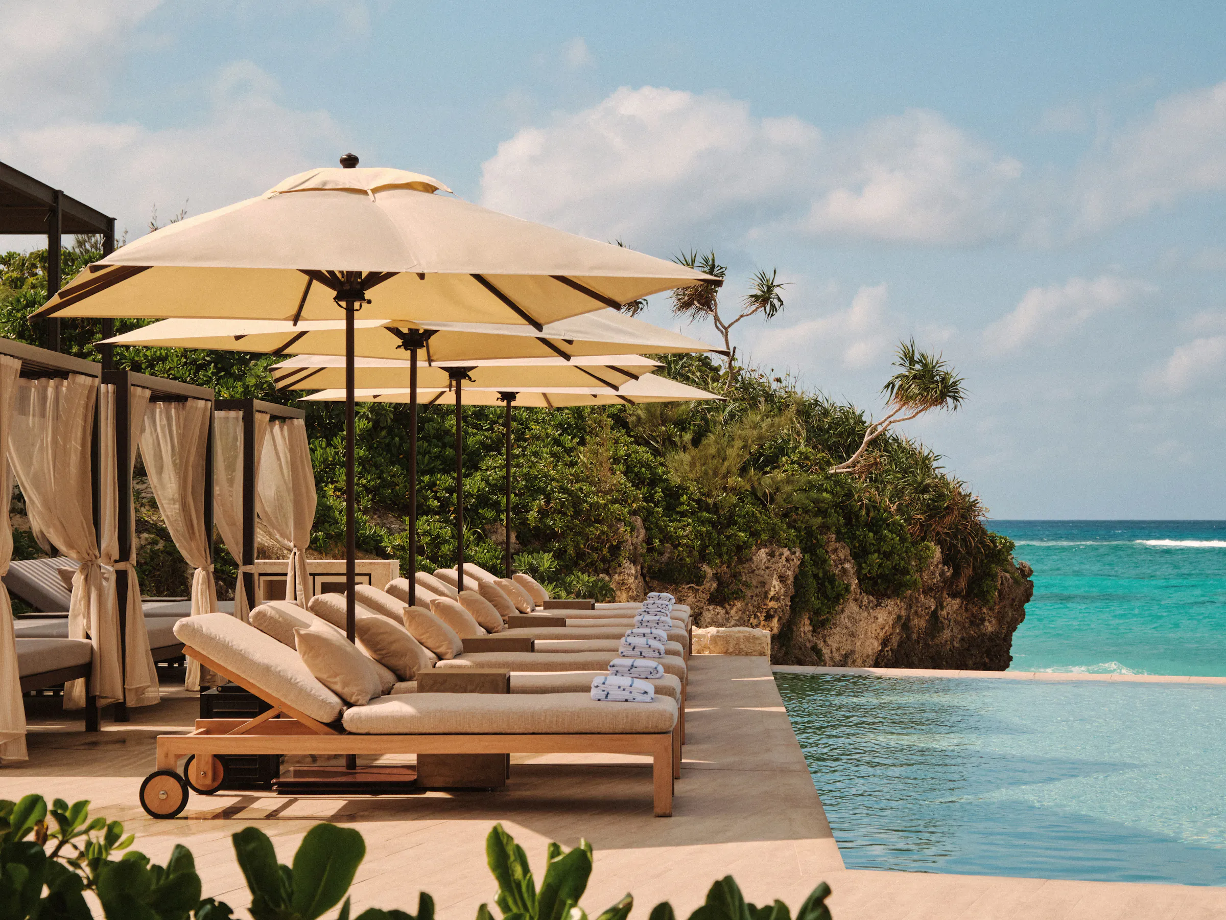 Cream loungers and umbrellas line an infinity pool at Rosewood Miyakojima, overlooking bright blue coastal water.