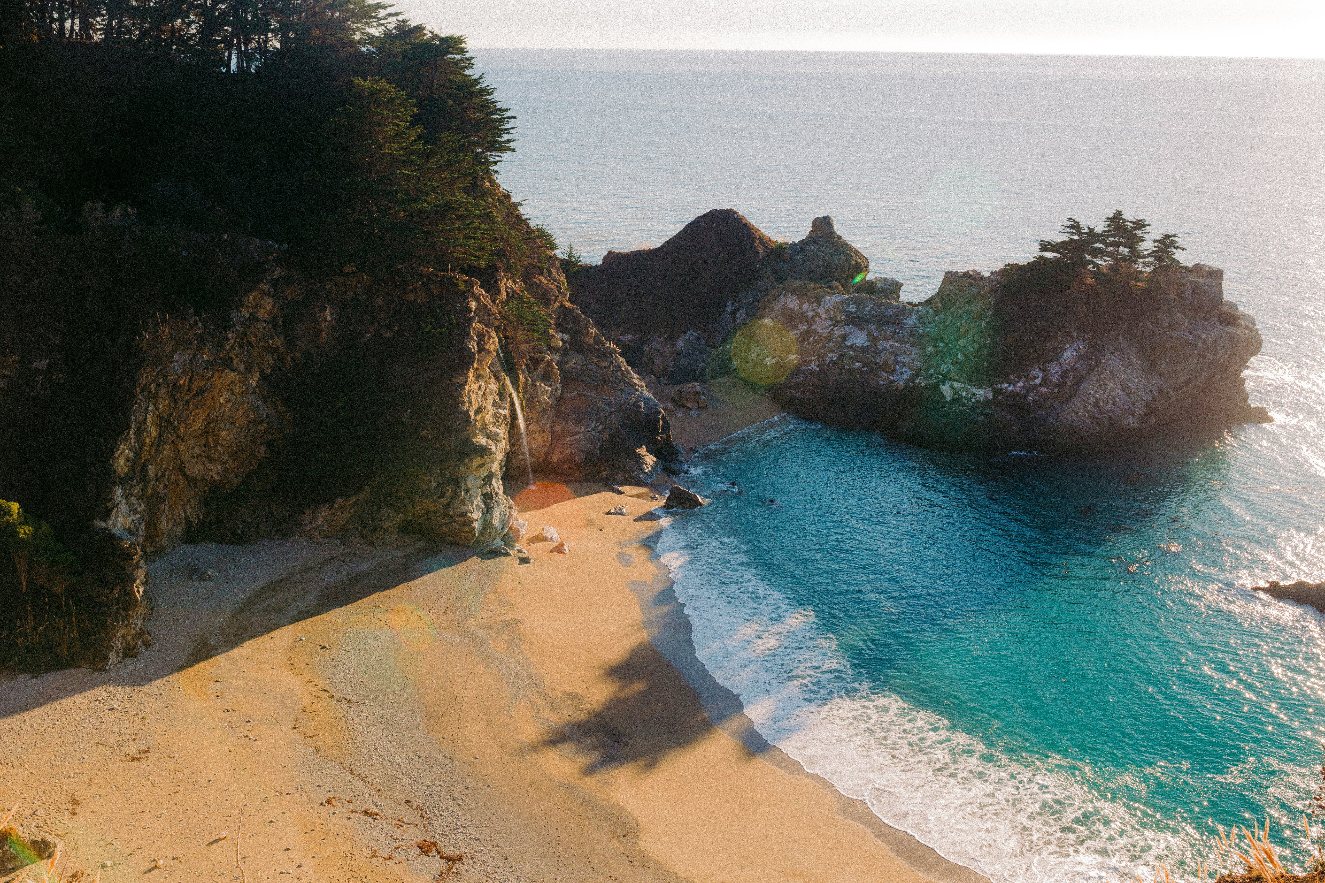 A secluded sandy cove curves beneath steep rocky cliffs in Okinawa, where turquoise waves wash onto the shore.