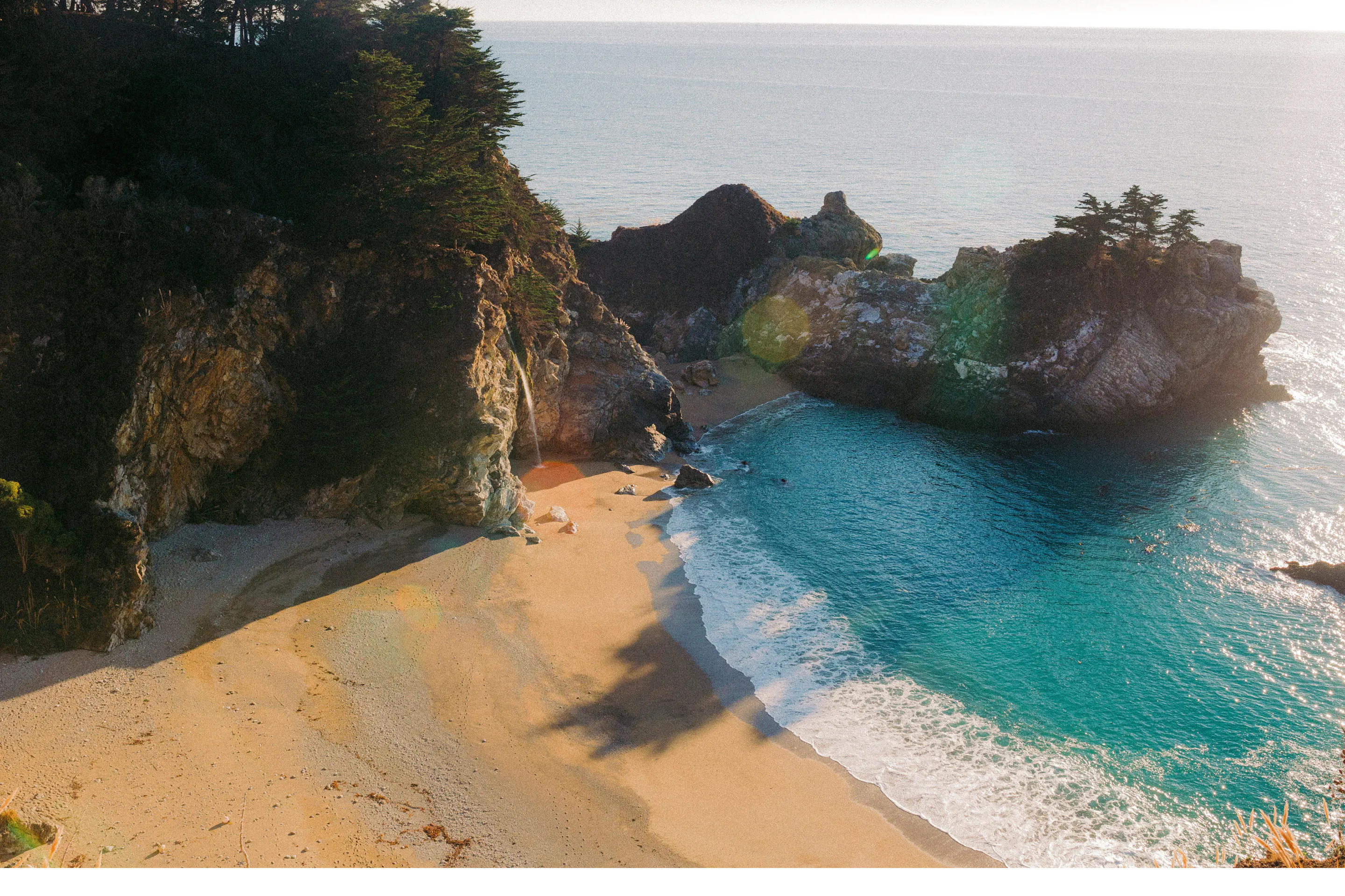 A secluded sandy cove curves beneath steep rocky cliffs in Okinawa, where turquoise waves wash onto the shore.