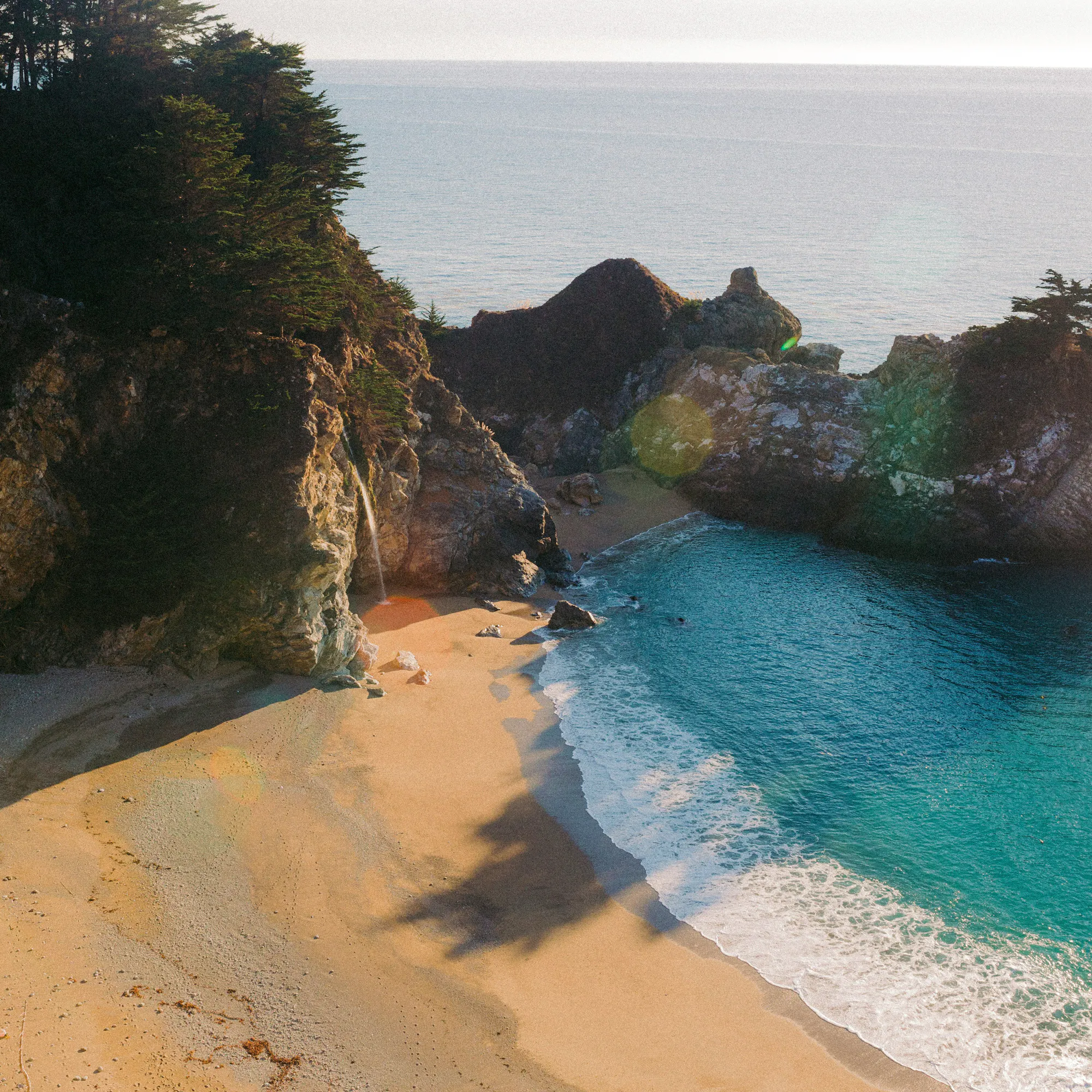 A secluded sandy cove curves beneath steep rocky cliffs in Okinawa, where turquoise waves wash onto the shore.