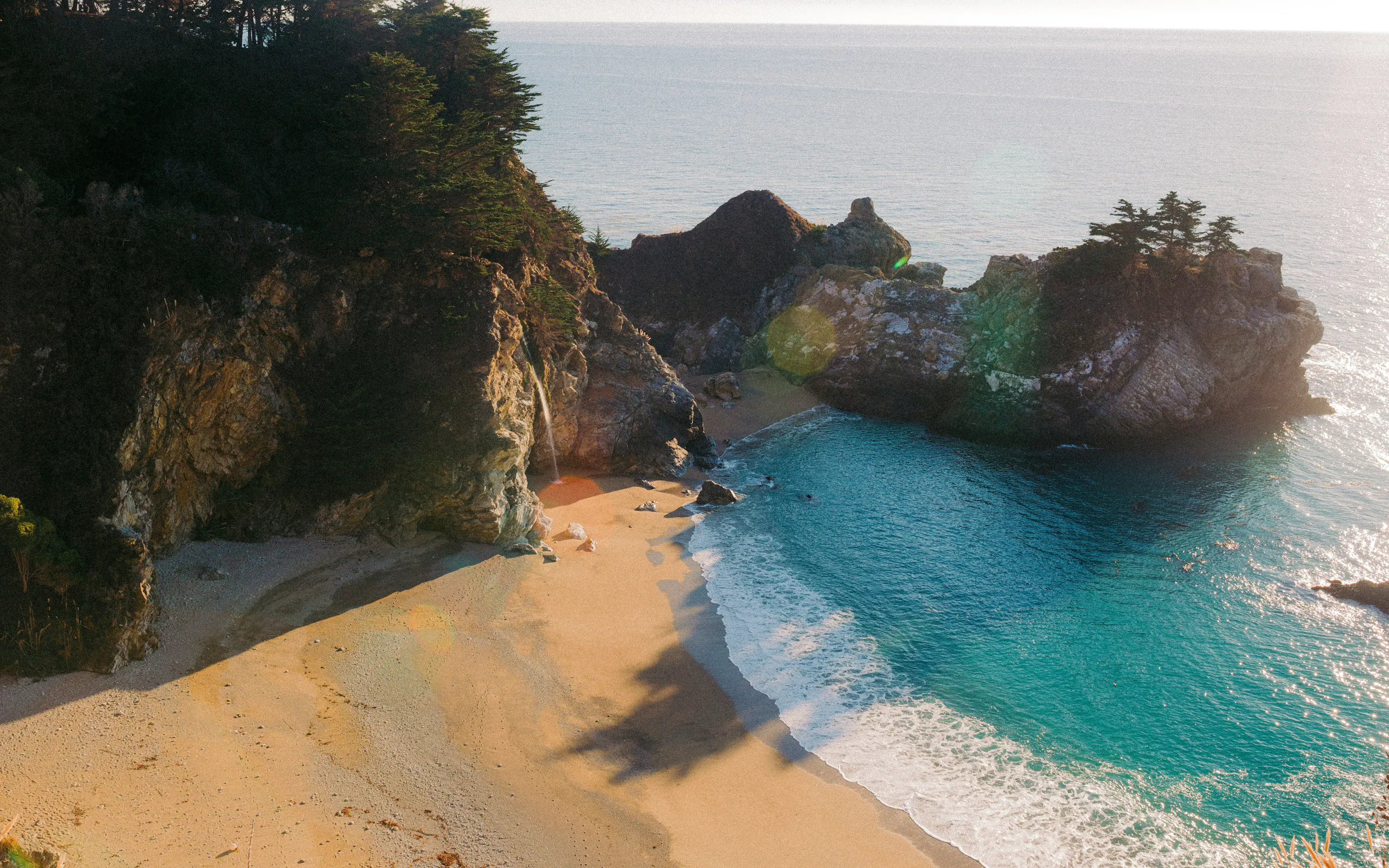 A secluded sandy cove curves beneath steep rocky cliffs in Okinawa, where turquoise waves wash onto the shore.
