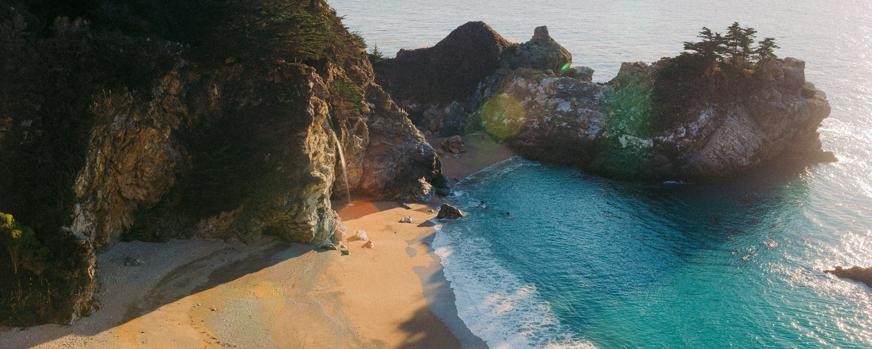 A secluded sandy cove curves beneath steep rocky cliffs in Okinawa, where turquoise waves wash onto the shore.