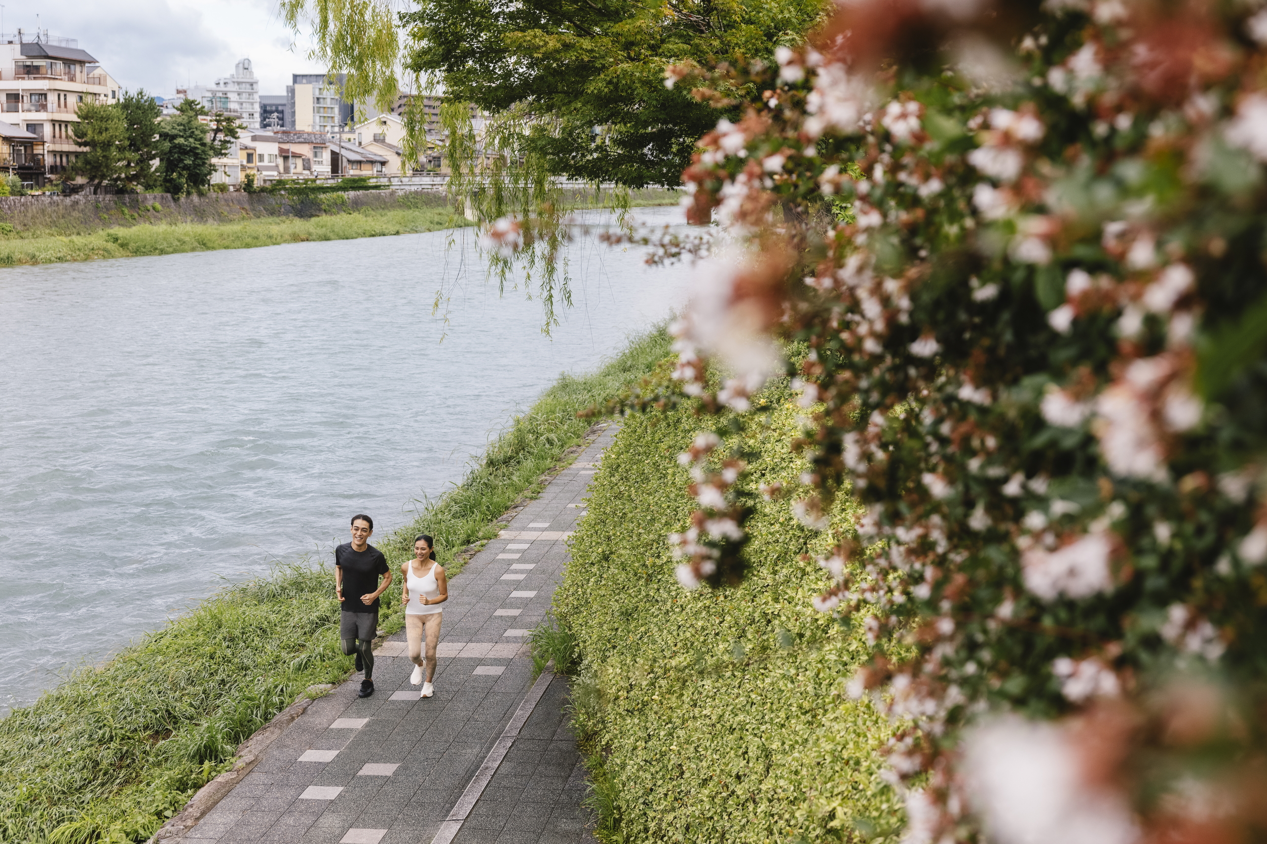 Two runners move along a riverside path in Kyoto, with spring blossoms in the foreground and city homes beyond.