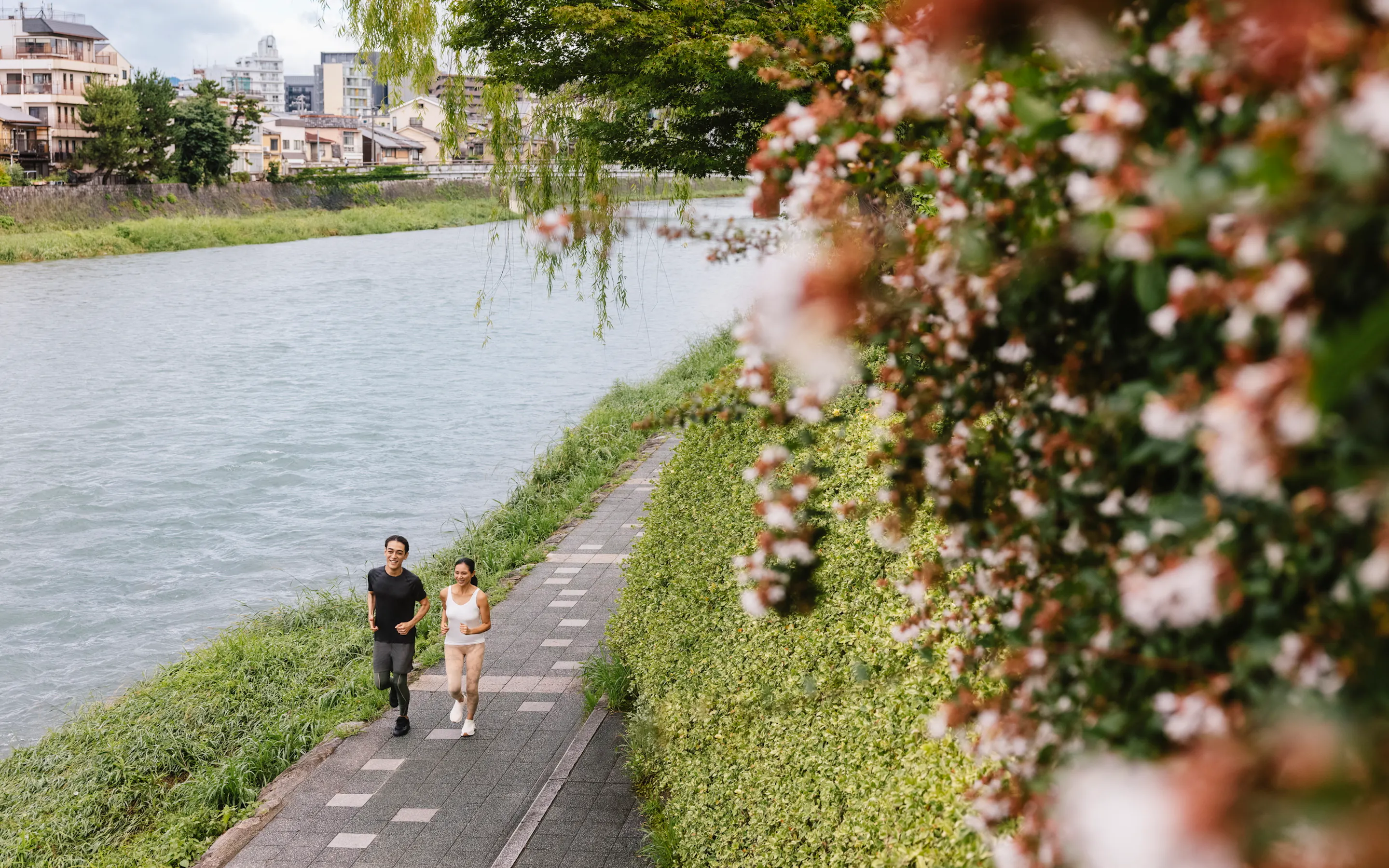 Two runners move along a riverside path in Kyoto, with spring blossoms in the foreground and city homes beyond.