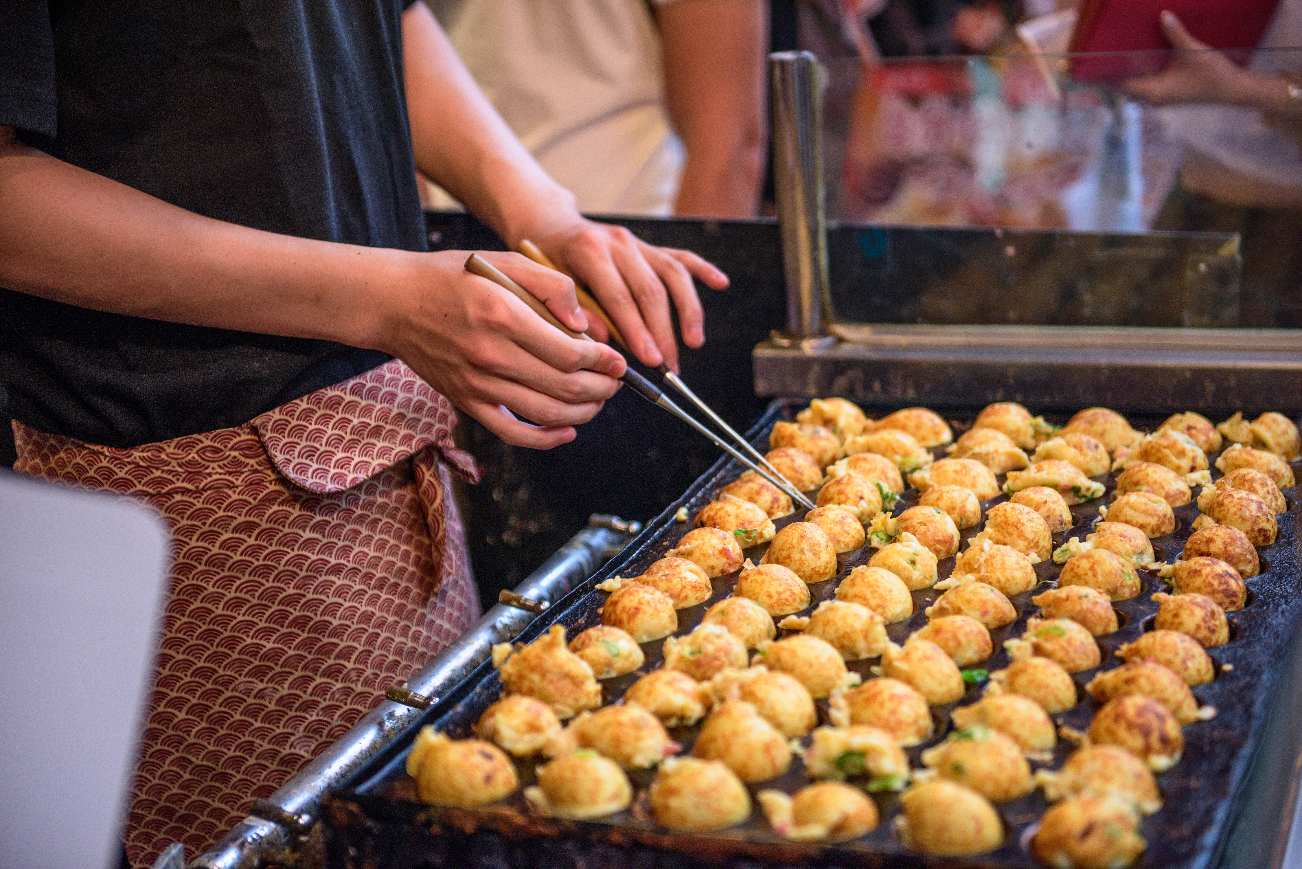 A street vendor turns rows of takoyaki on a hot griddle in Osaka, with diners and glowing lights behind the stall.