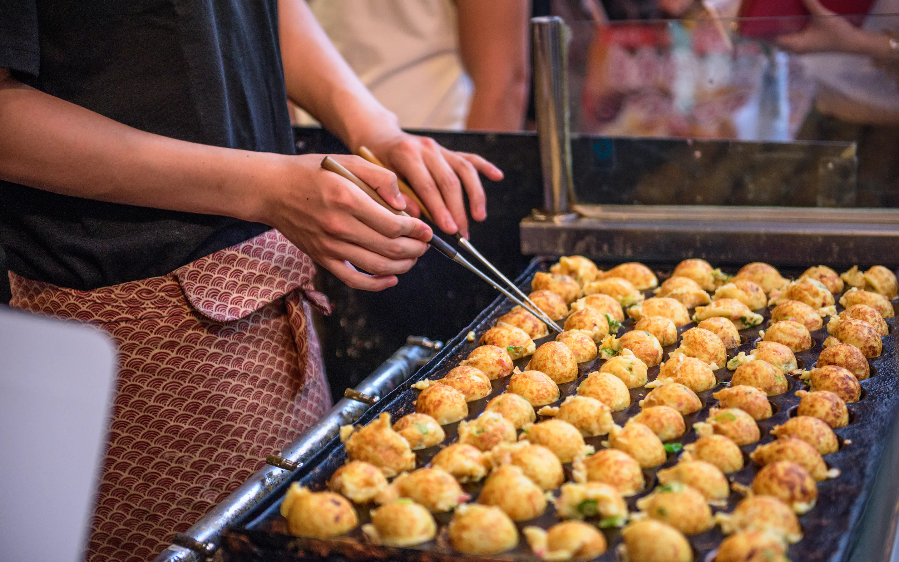 A street vendor turns rows of takoyaki on a hot griddle in Osaka, with diners and glowing lights behind the stall.