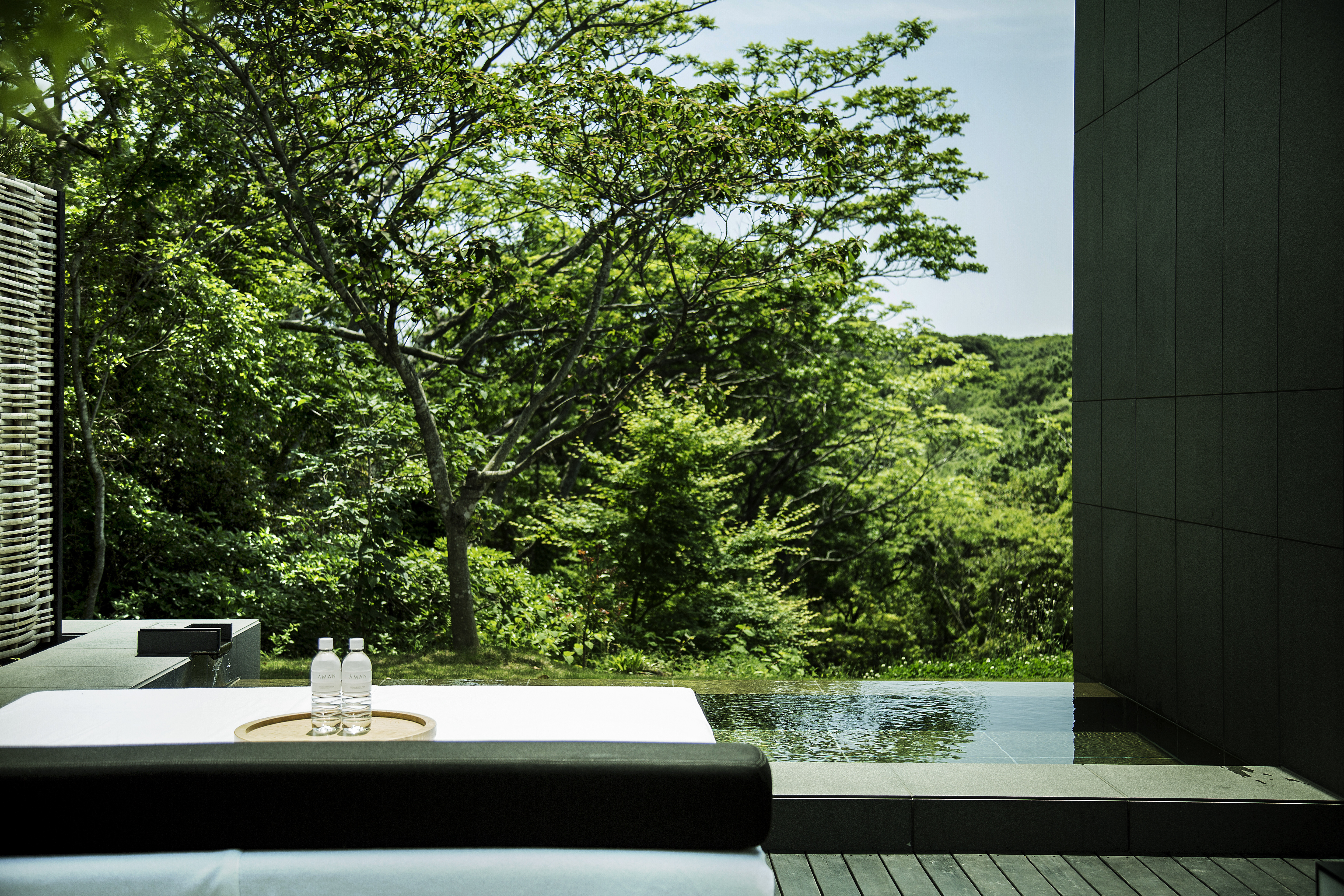 A private onsen terrace at Amanemu overlooks dense green trees, with still water, a cushioned bench, and two bottles.