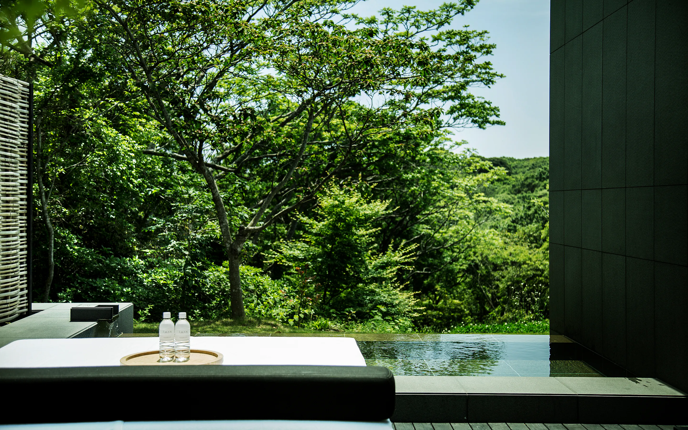 A private onsen terrace at Amanemu overlooks dense green trees, with still water, a cushioned bench, and two bottles.