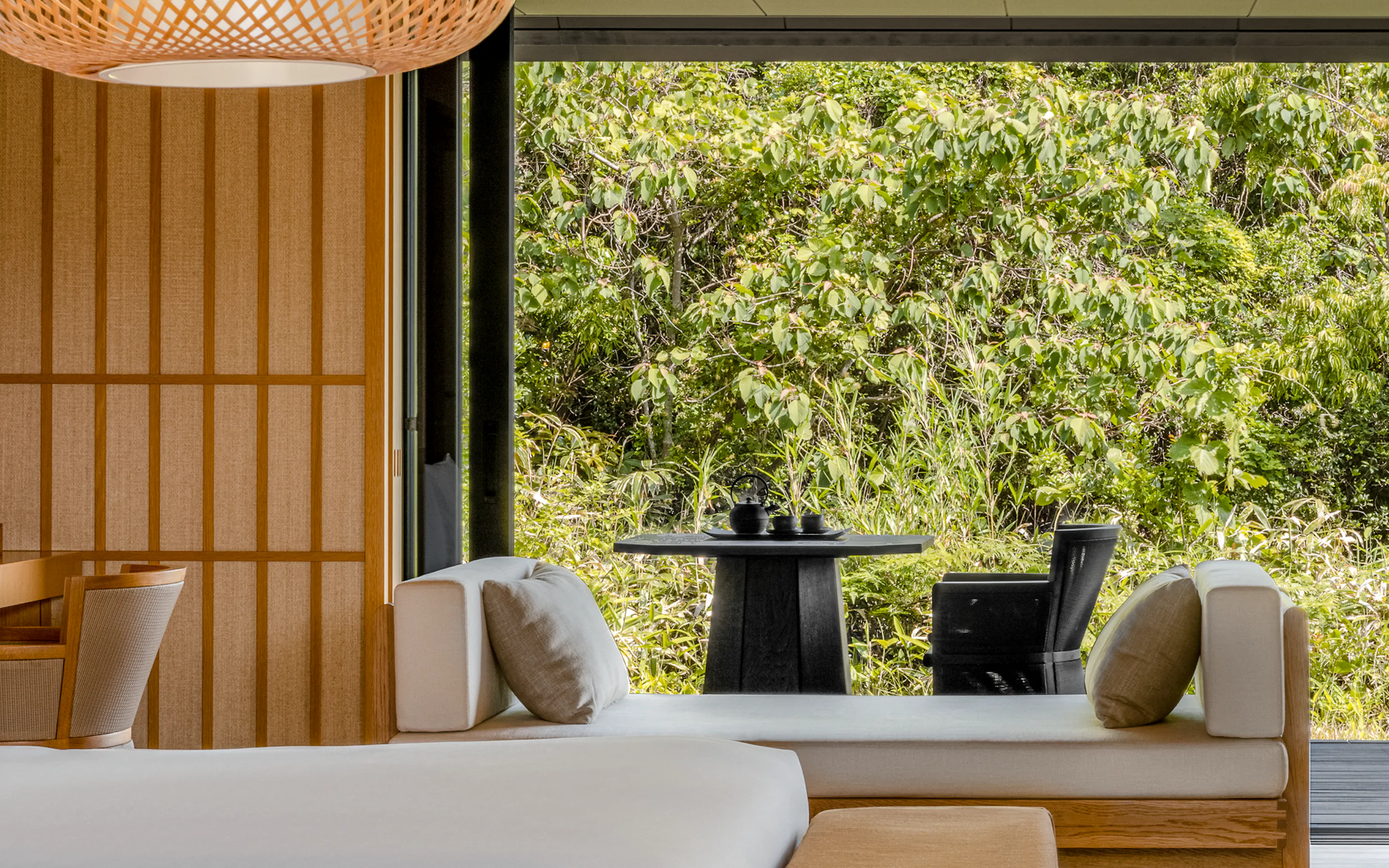 A bright Mori Suite bedroom at Amanemu opens to a private deck, with timber walls, a daybed, and forest beyond.