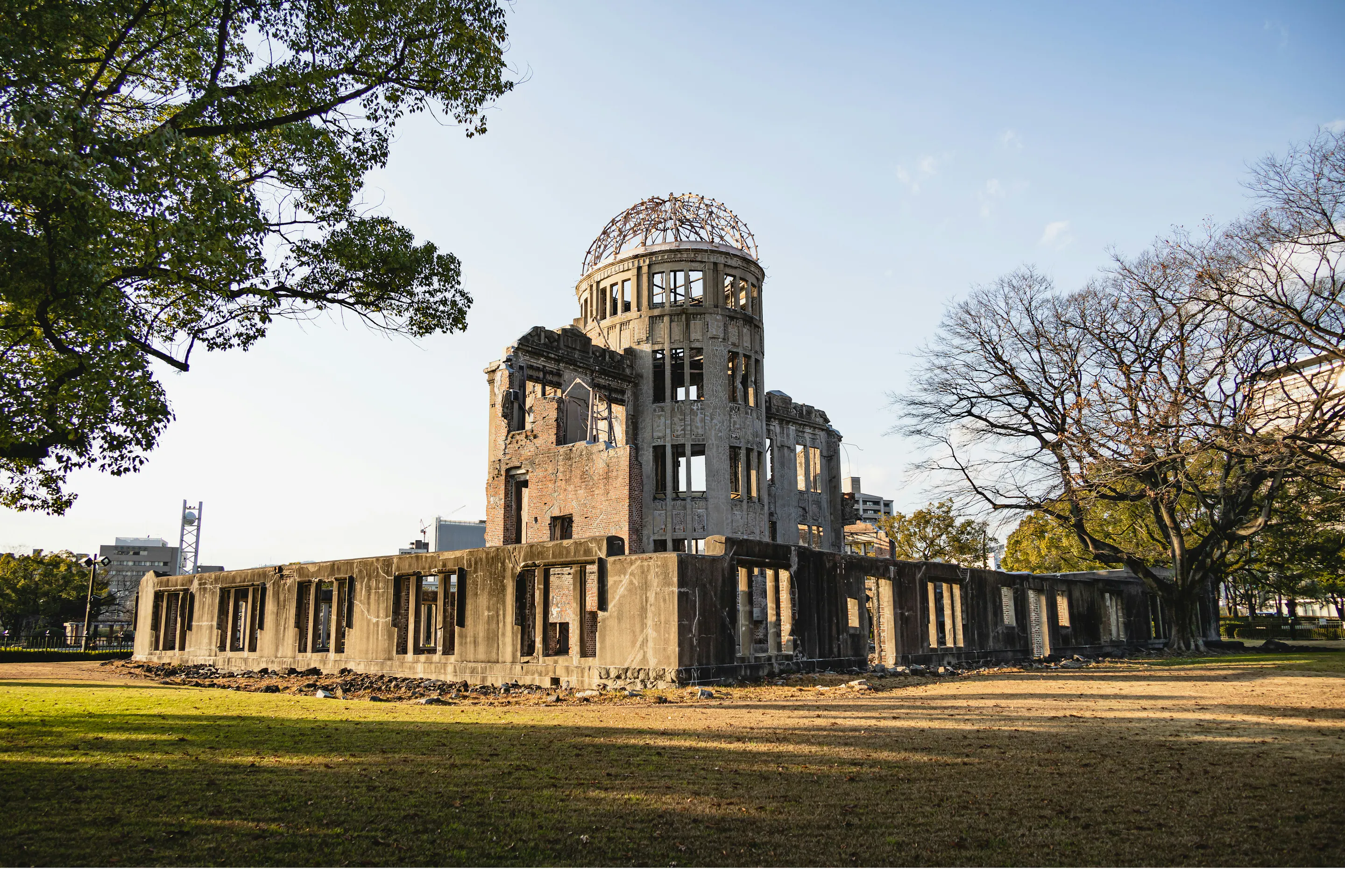 The Atomic Bomb Dome stands in warm light above the lawn, framed by broad trees and the surviving concrete walls.