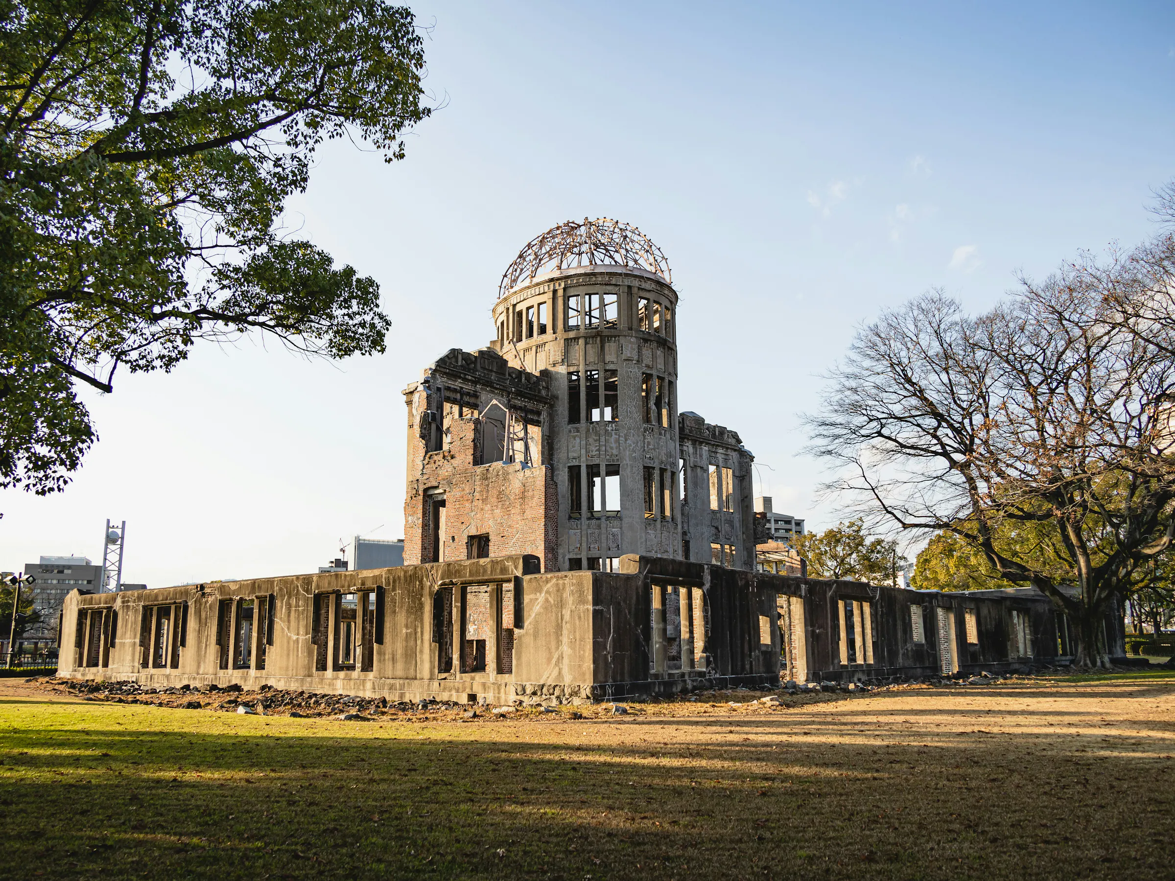 The Atomic Bomb Dome stands in warm light above the lawn, framed by broad trees and the surviving concrete walls.