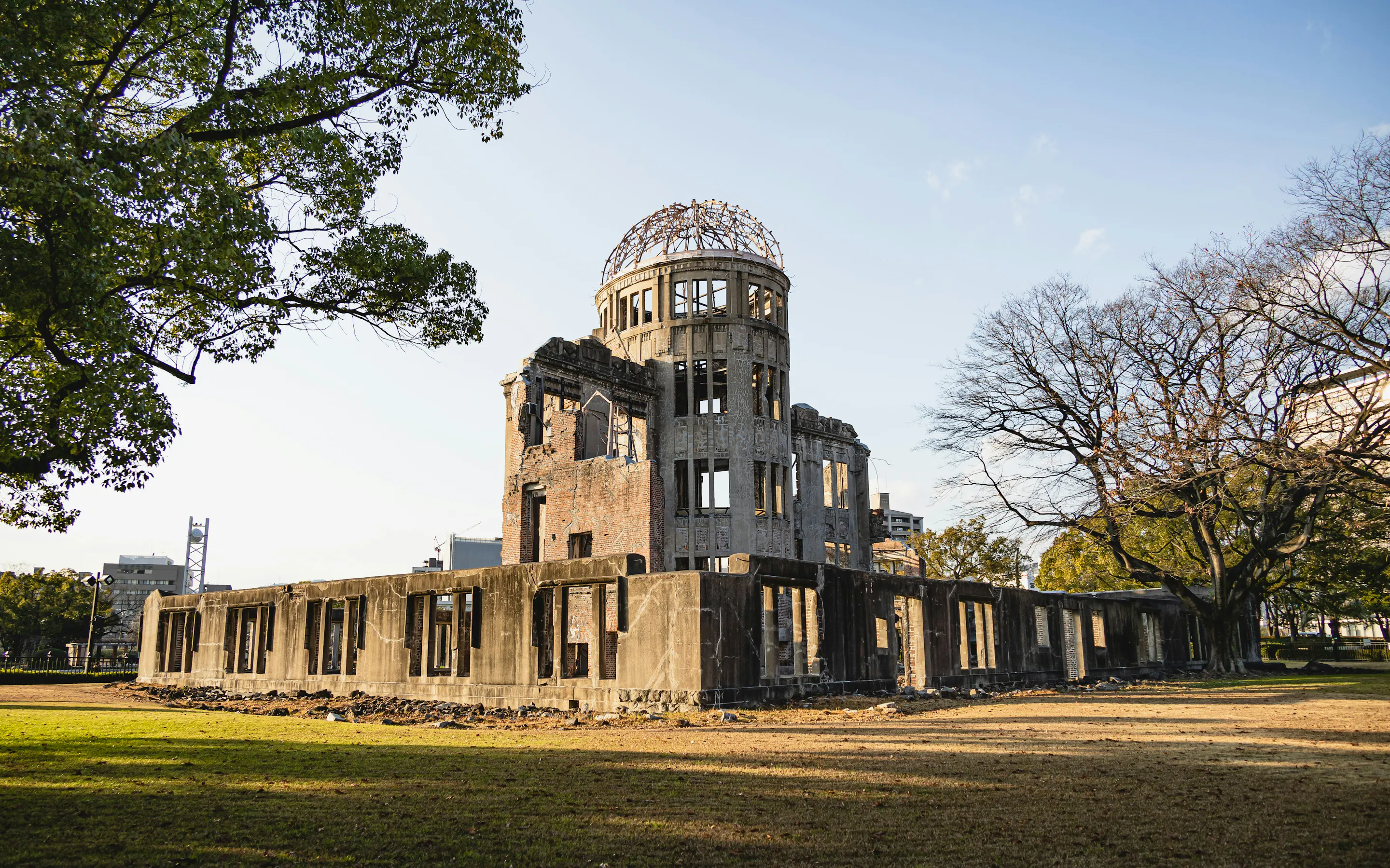 The Atomic Bomb Dome stands in warm light above the lawn, framed by broad trees and the surviving concrete walls.