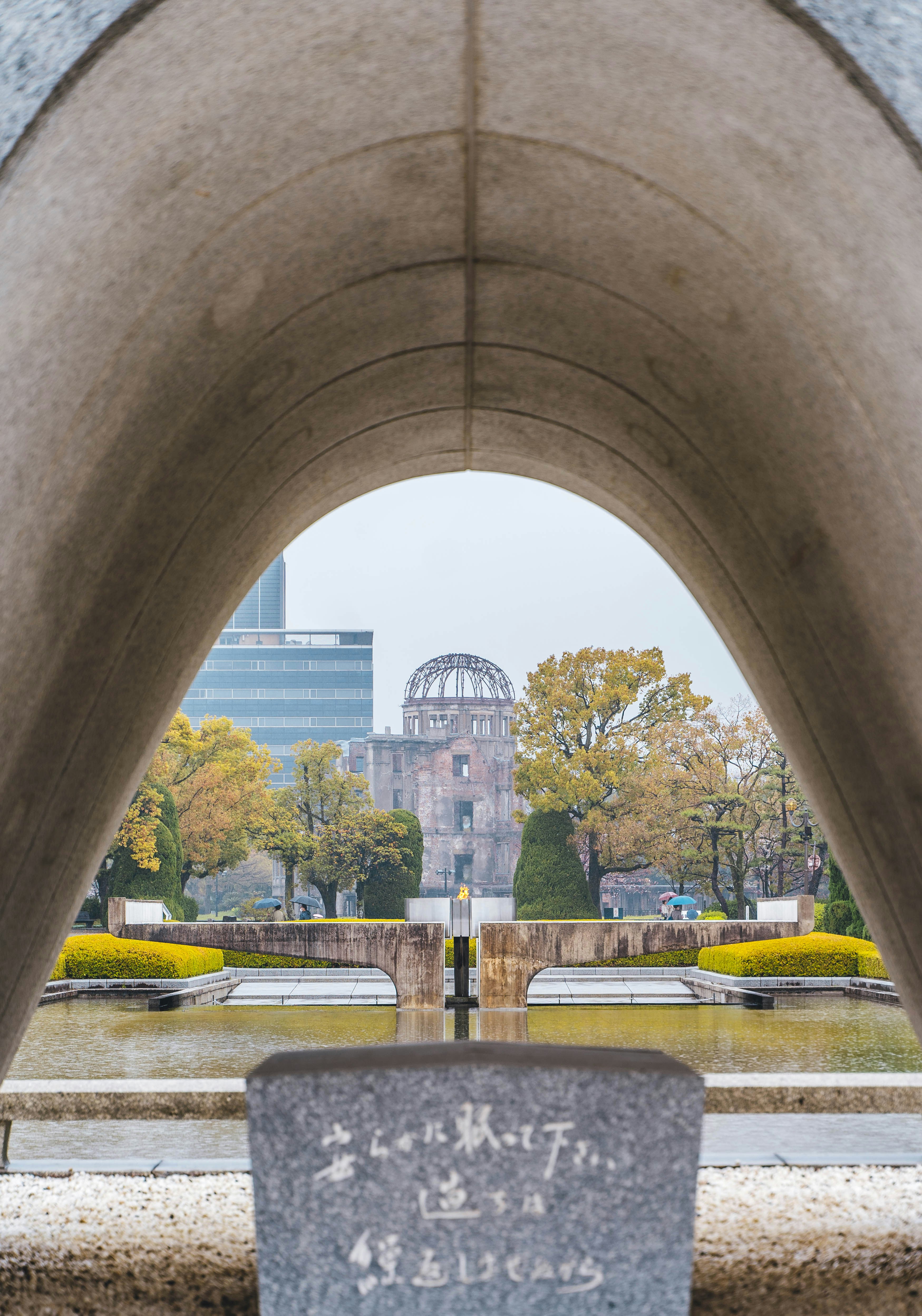 The Hiroshima Peace Memorial rises beyond the cenotaph arch, framed by still water, clipped hedges, and autumn trees.