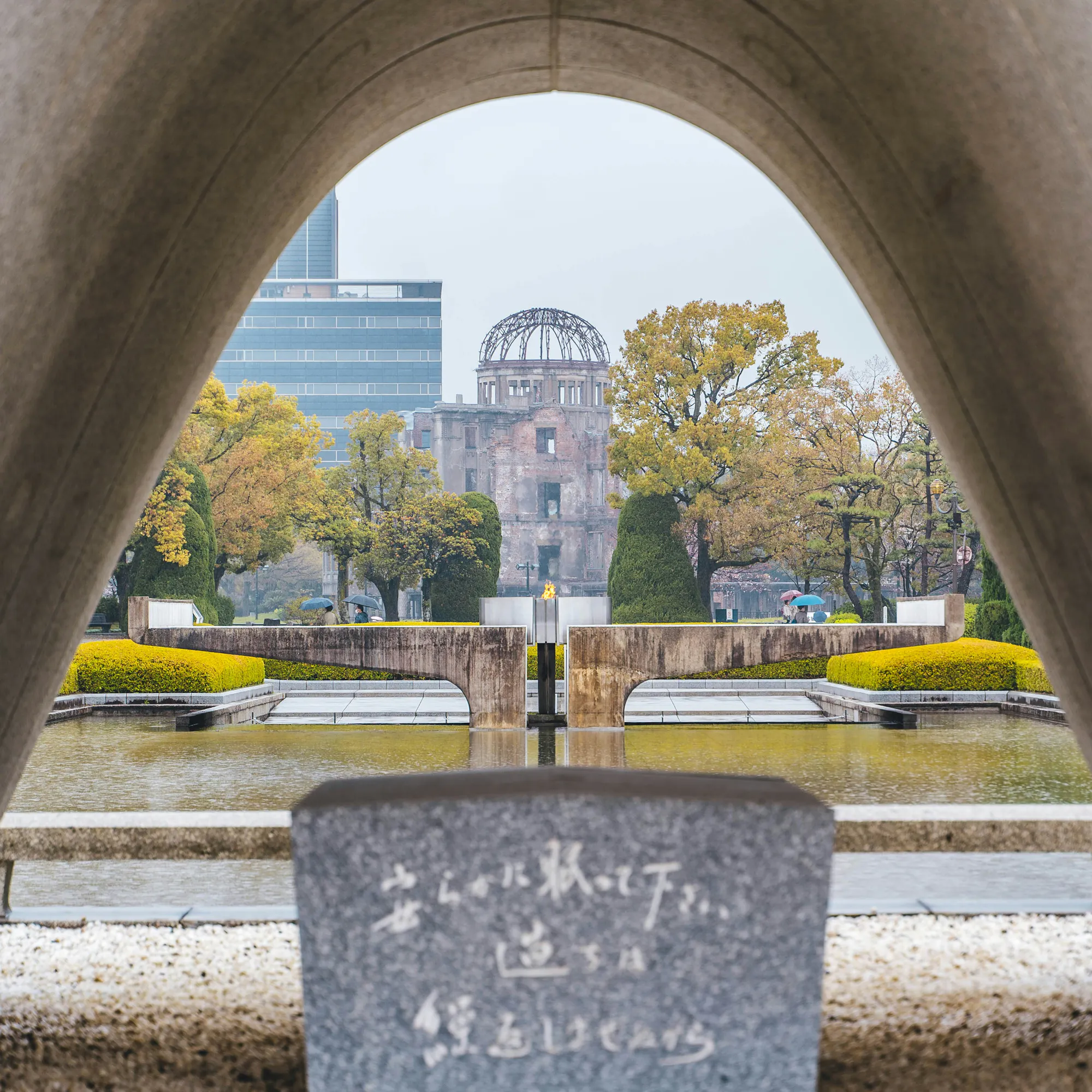 The Hiroshima Peace Memorial rises beyond the cenotaph arch, framed by still water, clipped hedges, and autumn trees.
