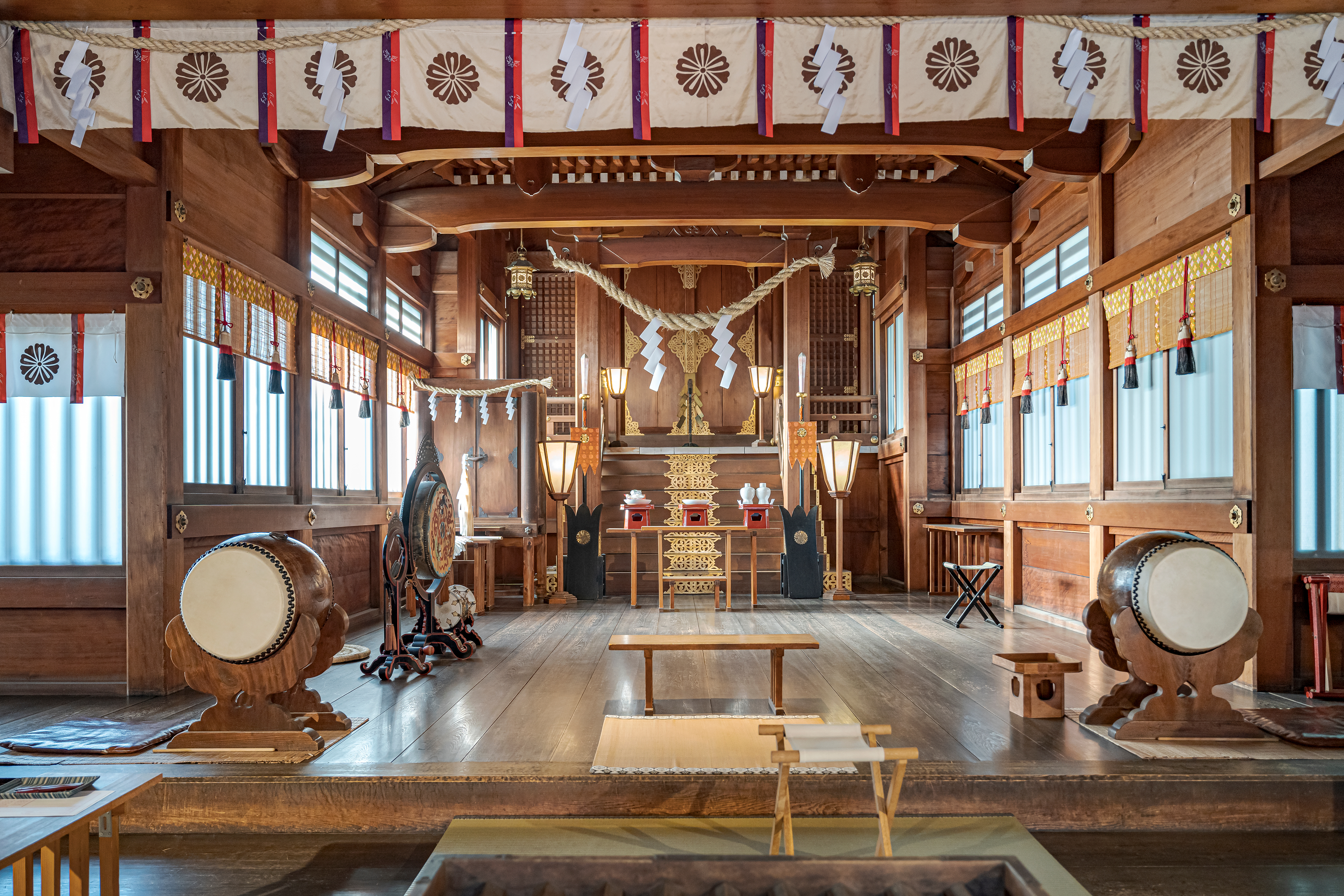 Lanterns, drums, and an altar line a wooden shrine hall, with warm daylight falling across the polished floor.