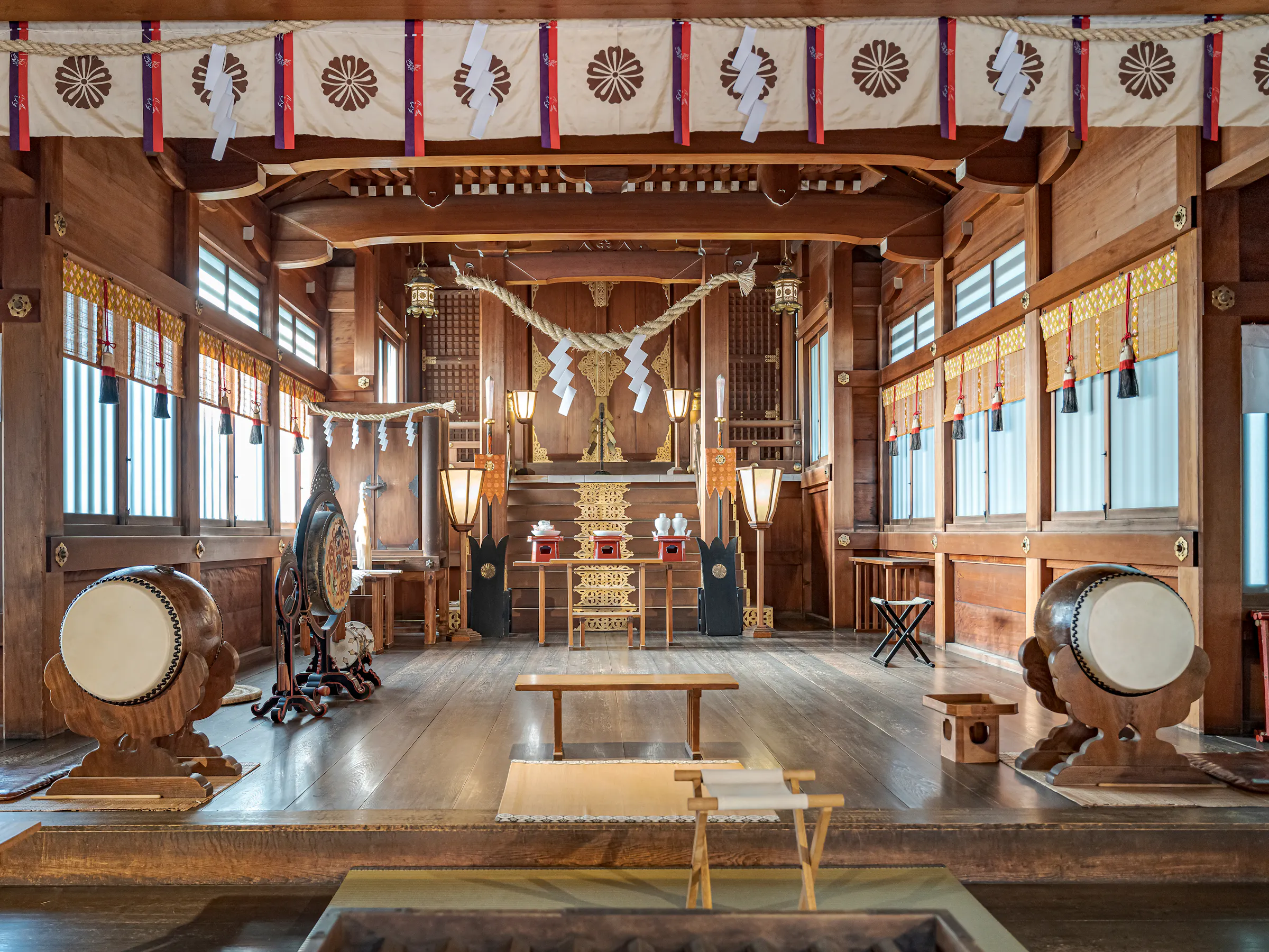 Lanterns, drums, and an altar line a wooden shrine hall, with warm daylight falling across the polished floor.