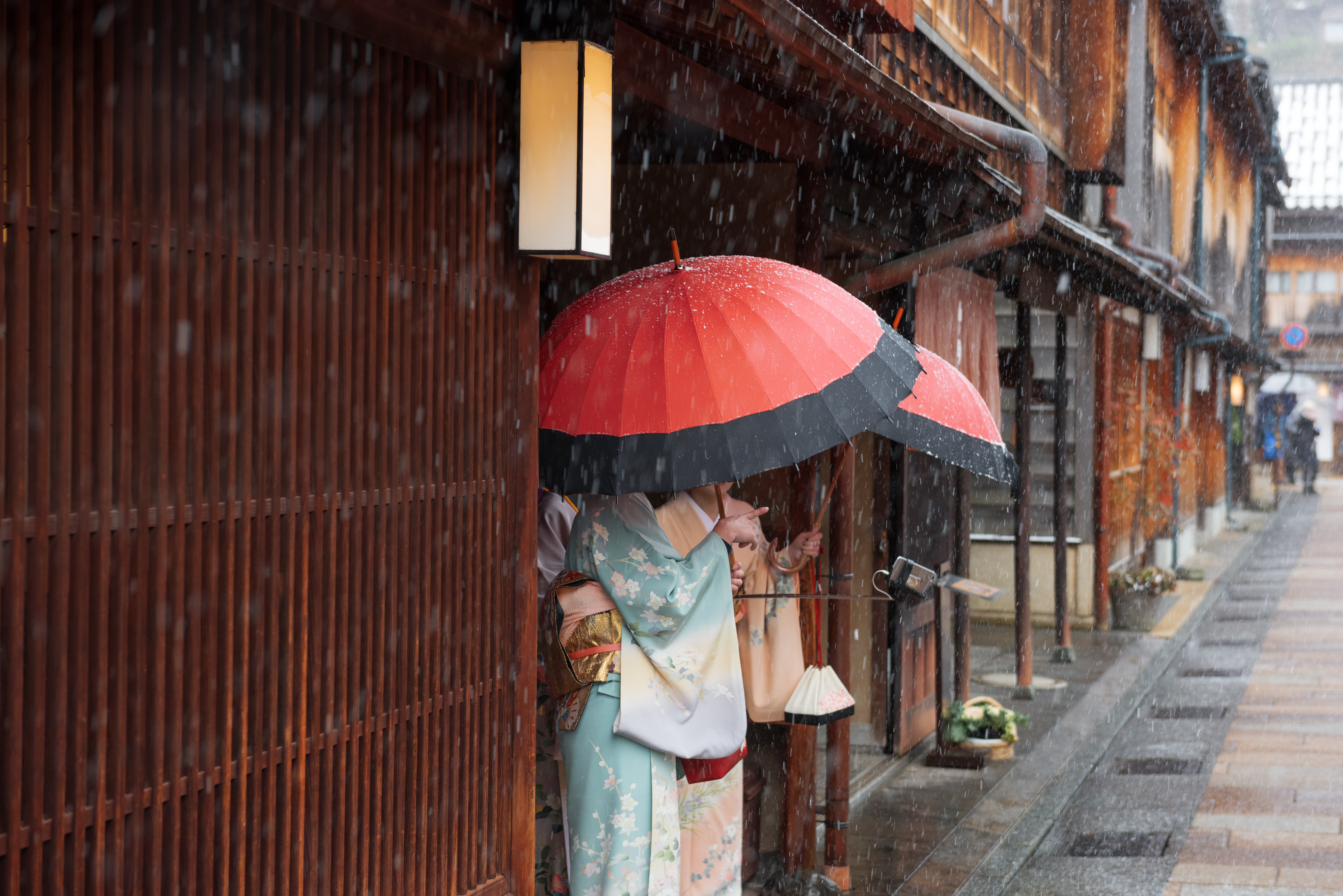 A person in a kimono stands under a red umbrella beside wooden facades as rain falls along a narrow old street.