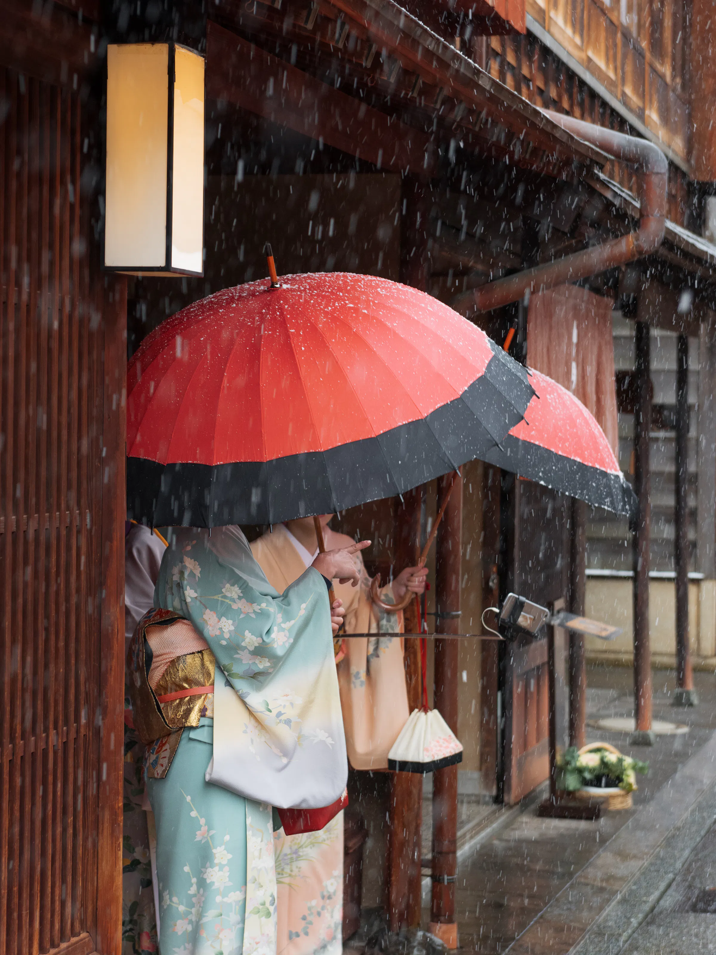 A person in a kimono stands under a red umbrella beside wooden facades as rain falls along a narrow old street.