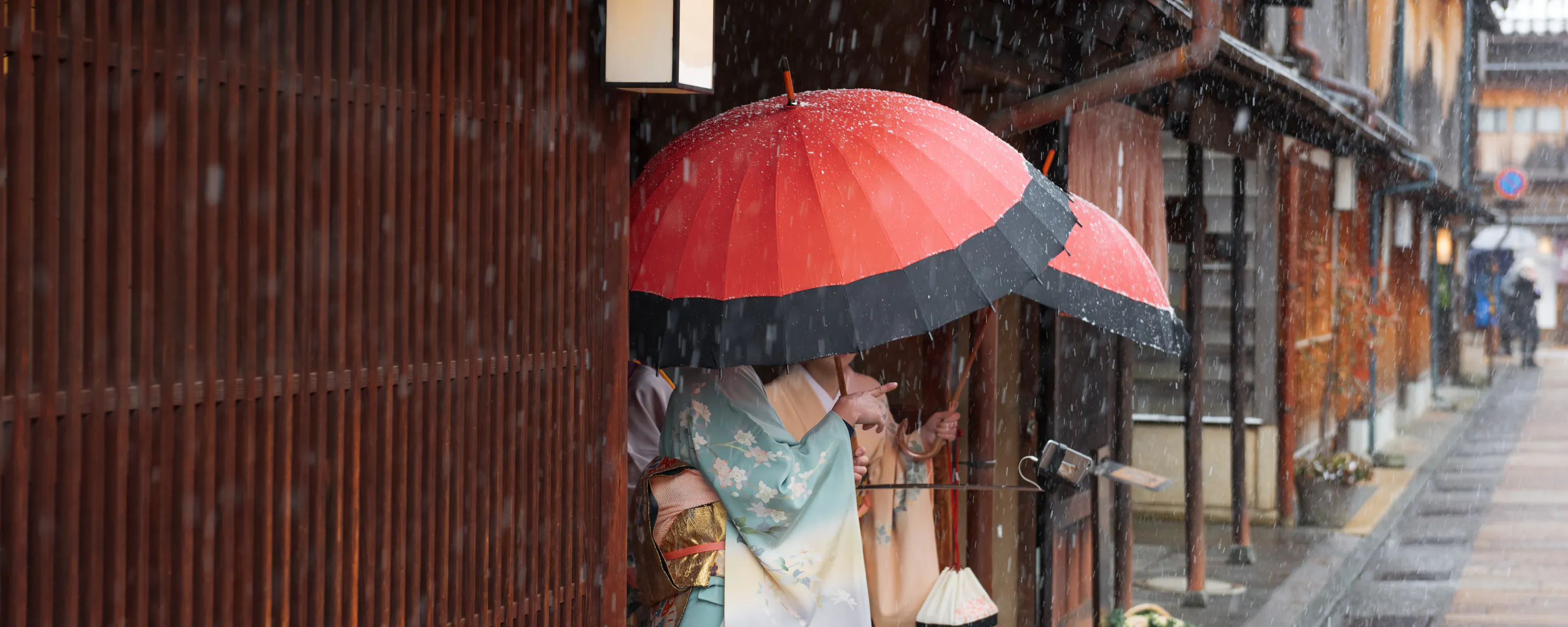 A person in a kimono stands under a red umbrella beside wooden facades as rain falls along a narrow old street.