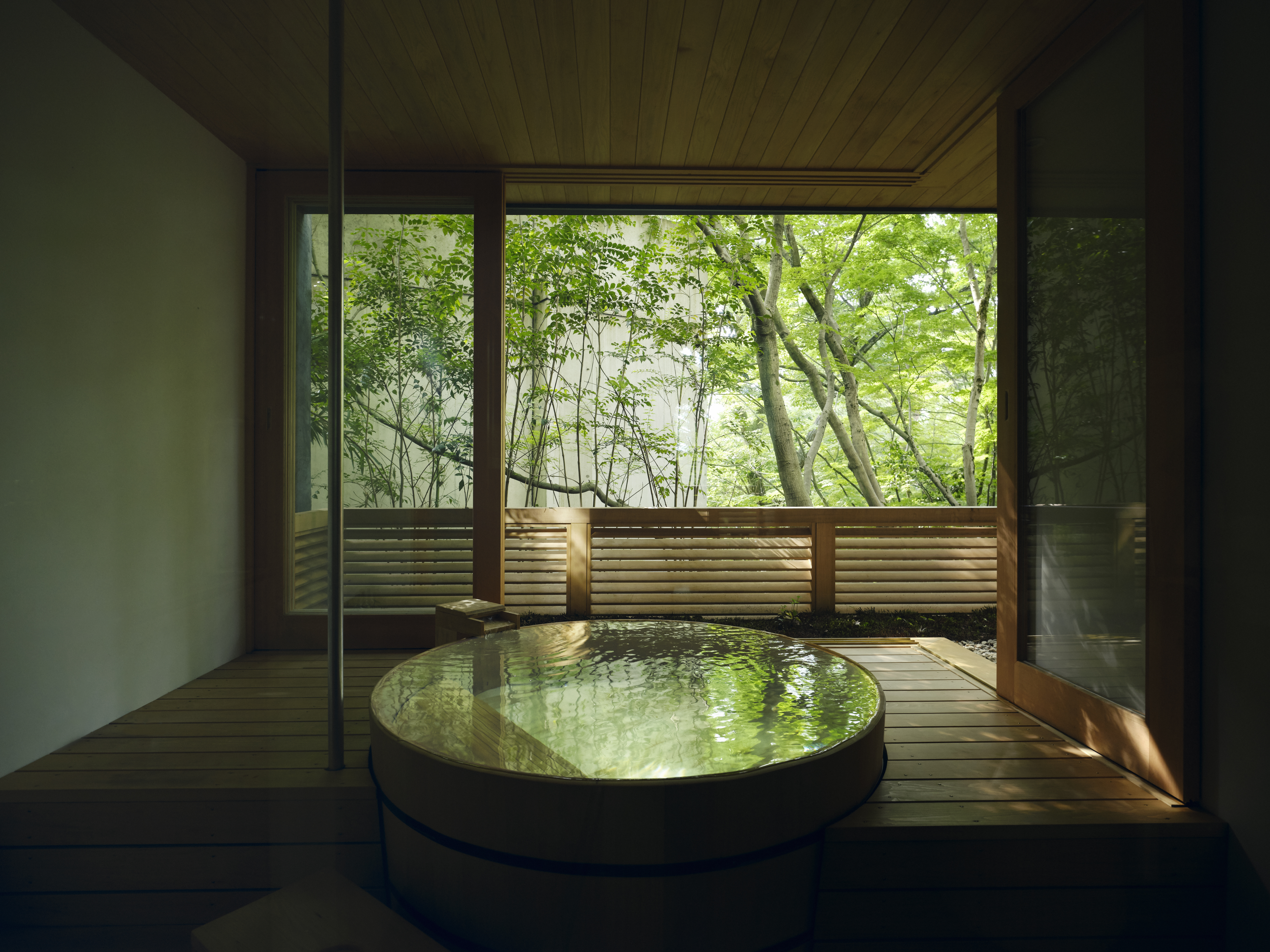 A round private onsen sits beside open sliding doors, with soft light filtering through green trees beyond the deck.