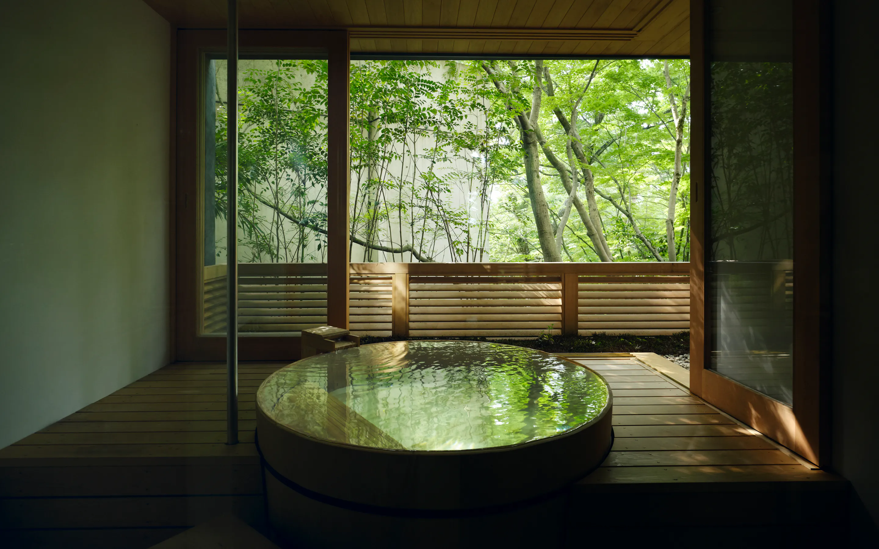 A round private onsen sits beside open sliding doors, with soft light filtering through green trees beyond the deck.