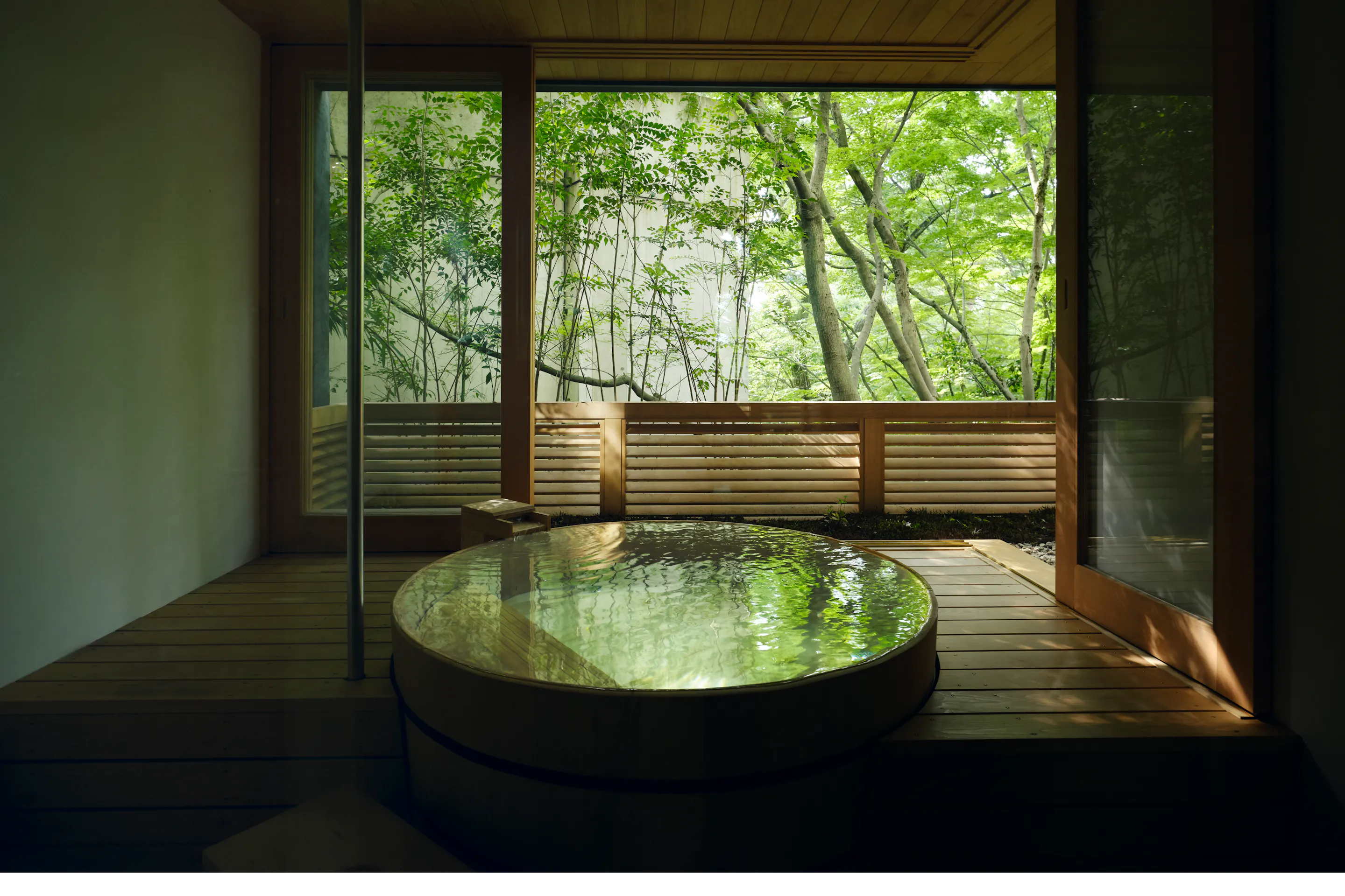 A round private onsen sits beside open sliding doors, with soft light filtering through green trees beyond the deck.