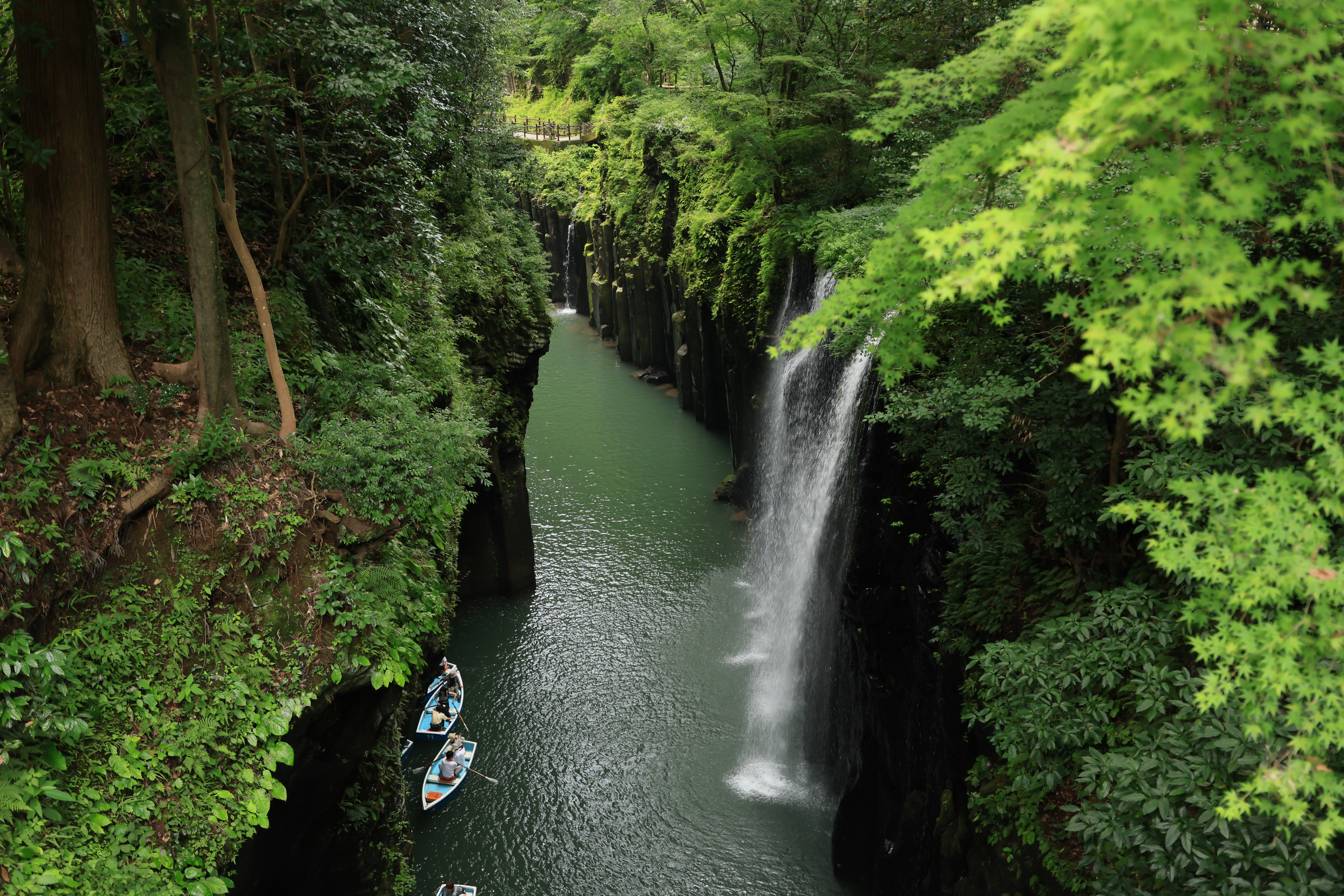 A narrow waterfall drops into a steep green gorge where small rowboats drift below towering basalt rock walls.