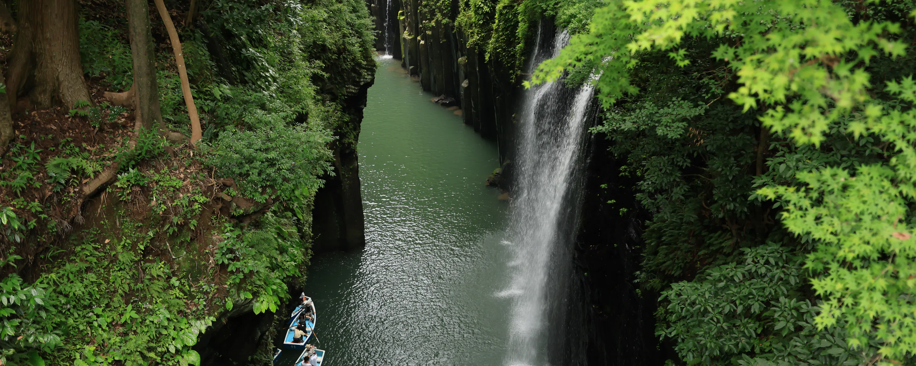 A narrow waterfall drops into a steep green gorge where small rowboats drift below towering basalt rock walls.