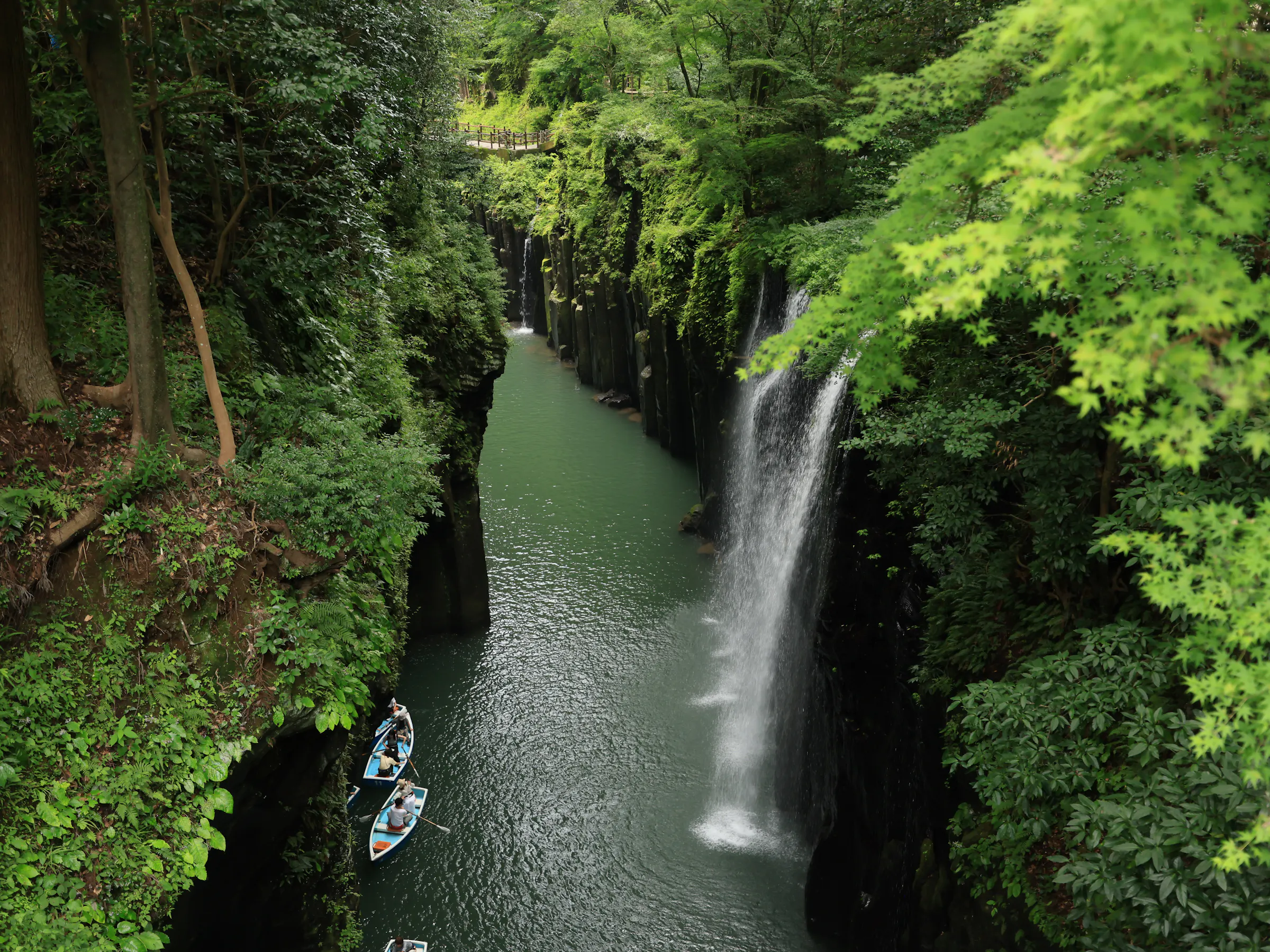 A narrow waterfall drops into a steep green gorge where small rowboats drift below towering basalt rock walls.