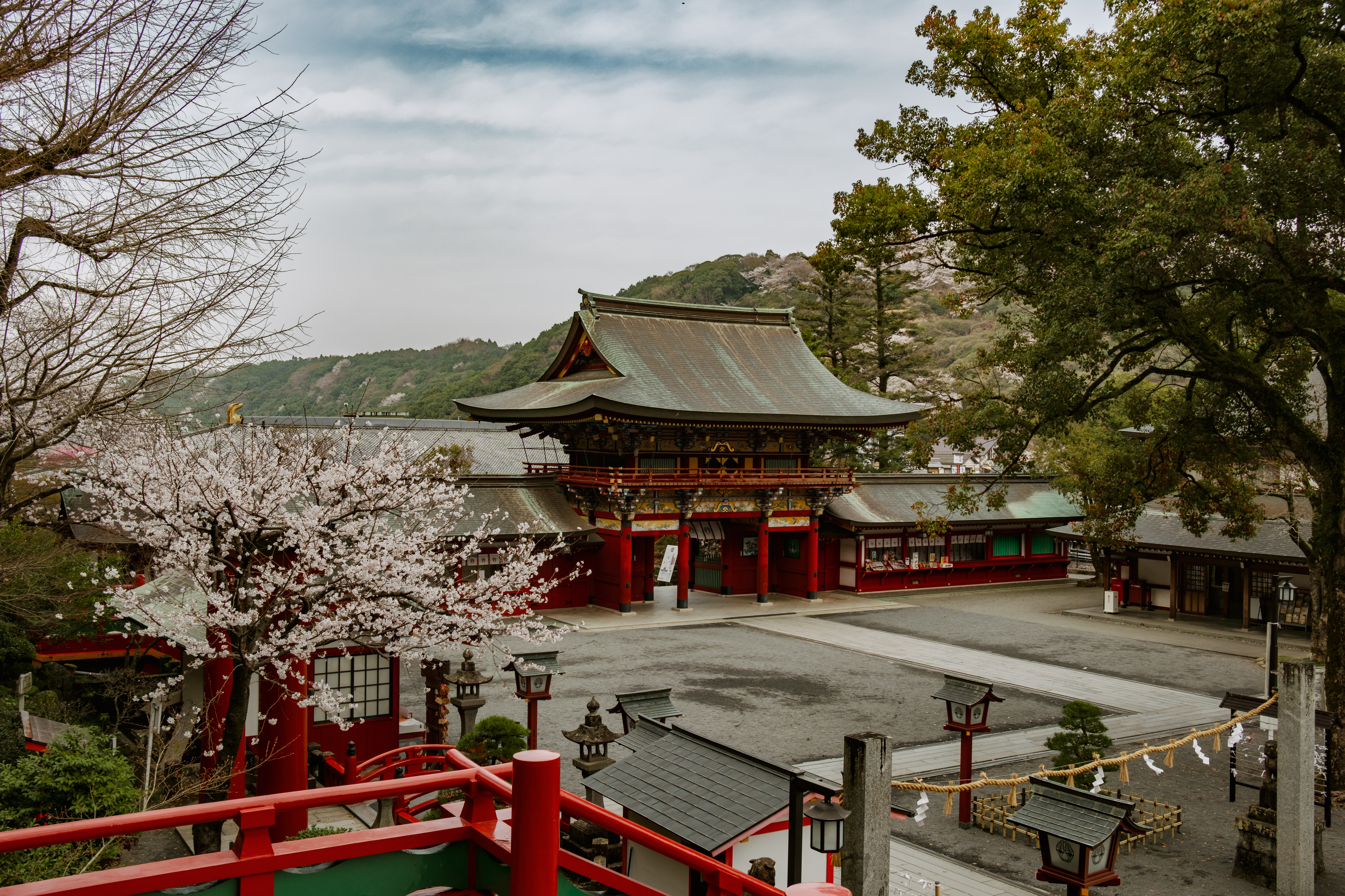 A red shrine building sits beneath pale cherry blossoms, with lanterns, stone markers, and wooded hills around it.
