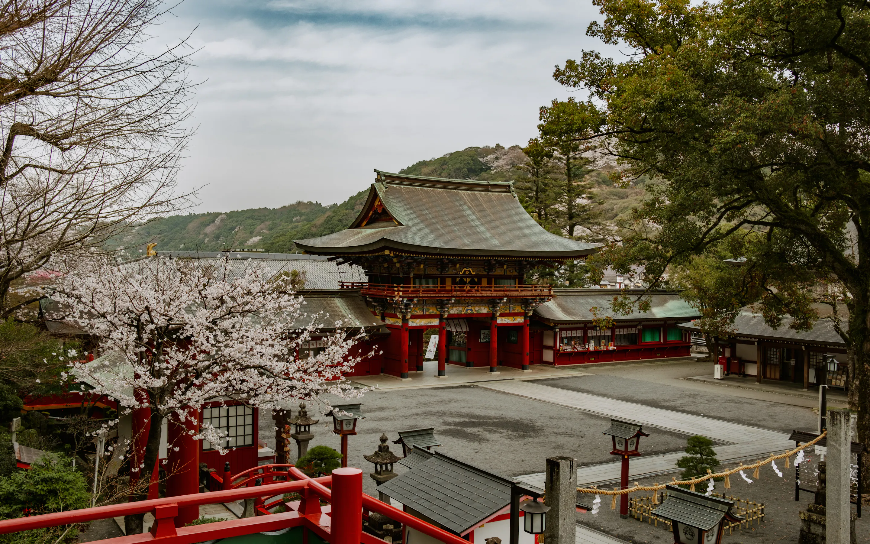 A red shrine building sits beneath pale cherry blossoms, with lanterns, stone markers, and wooded hills around it.