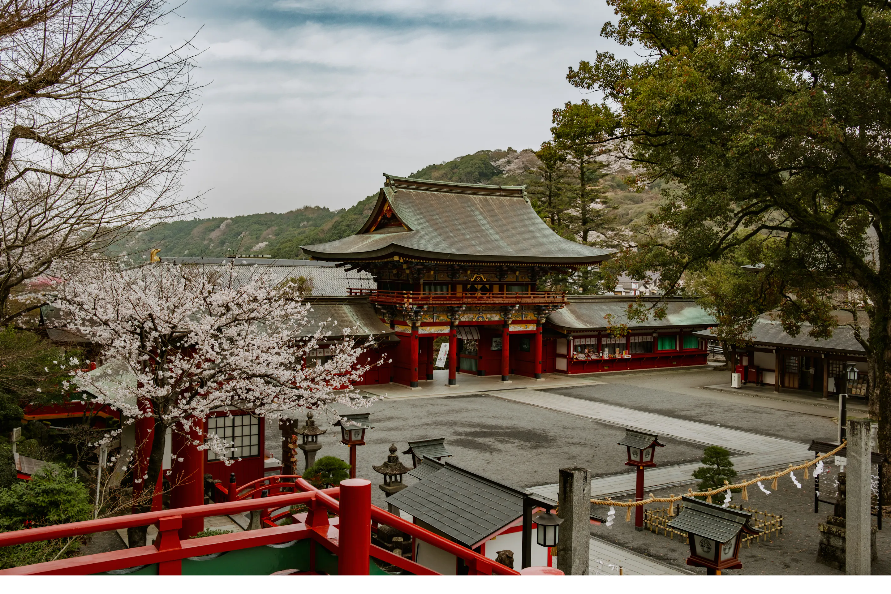 A red shrine building sits beneath pale cherry blossoms, with lanterns, stone markers, and wooded hills around it.