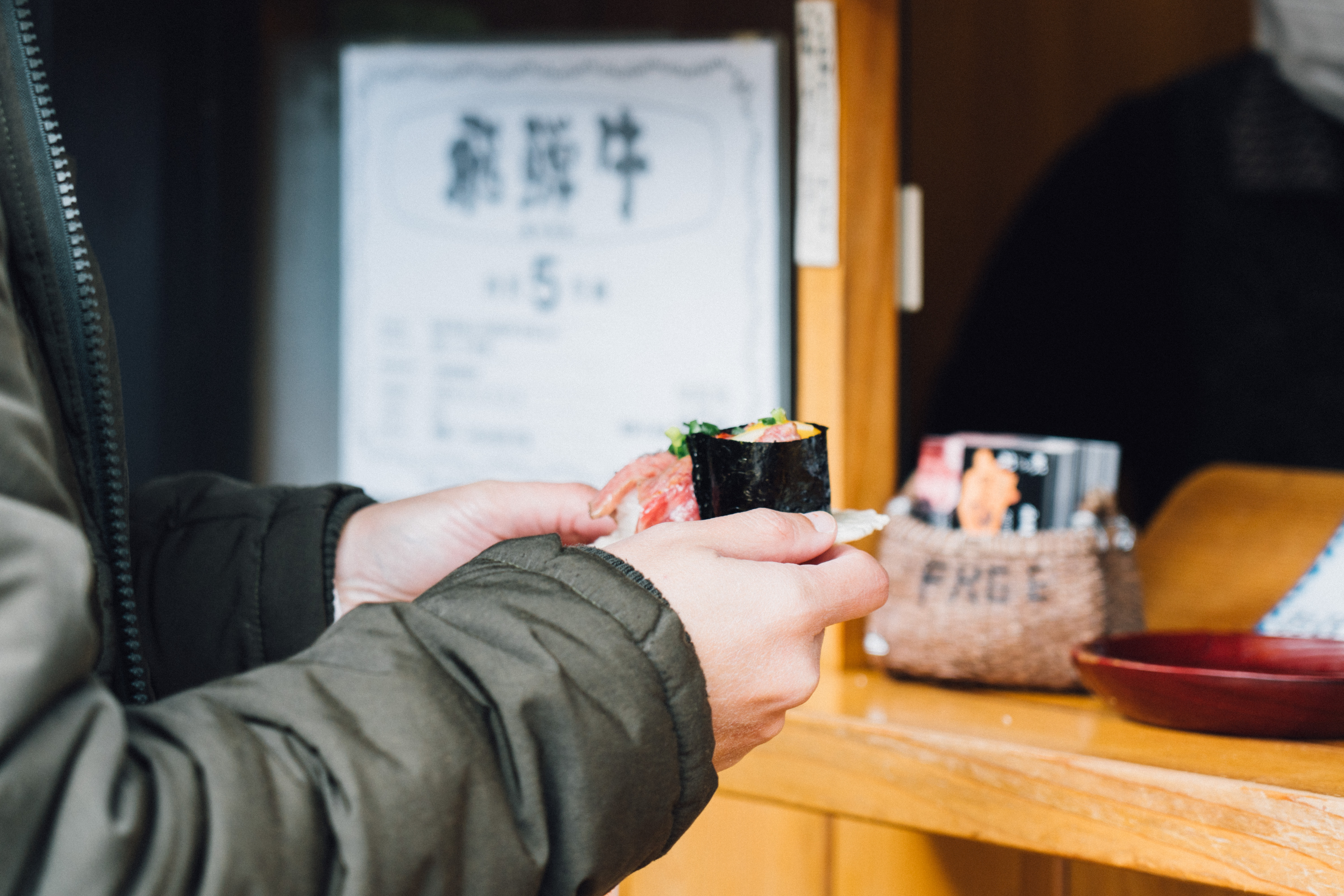 A diner holds a sushi hand roll at a wooden counter in Takayama, with small bowls, menus, and a blurred figure behind.