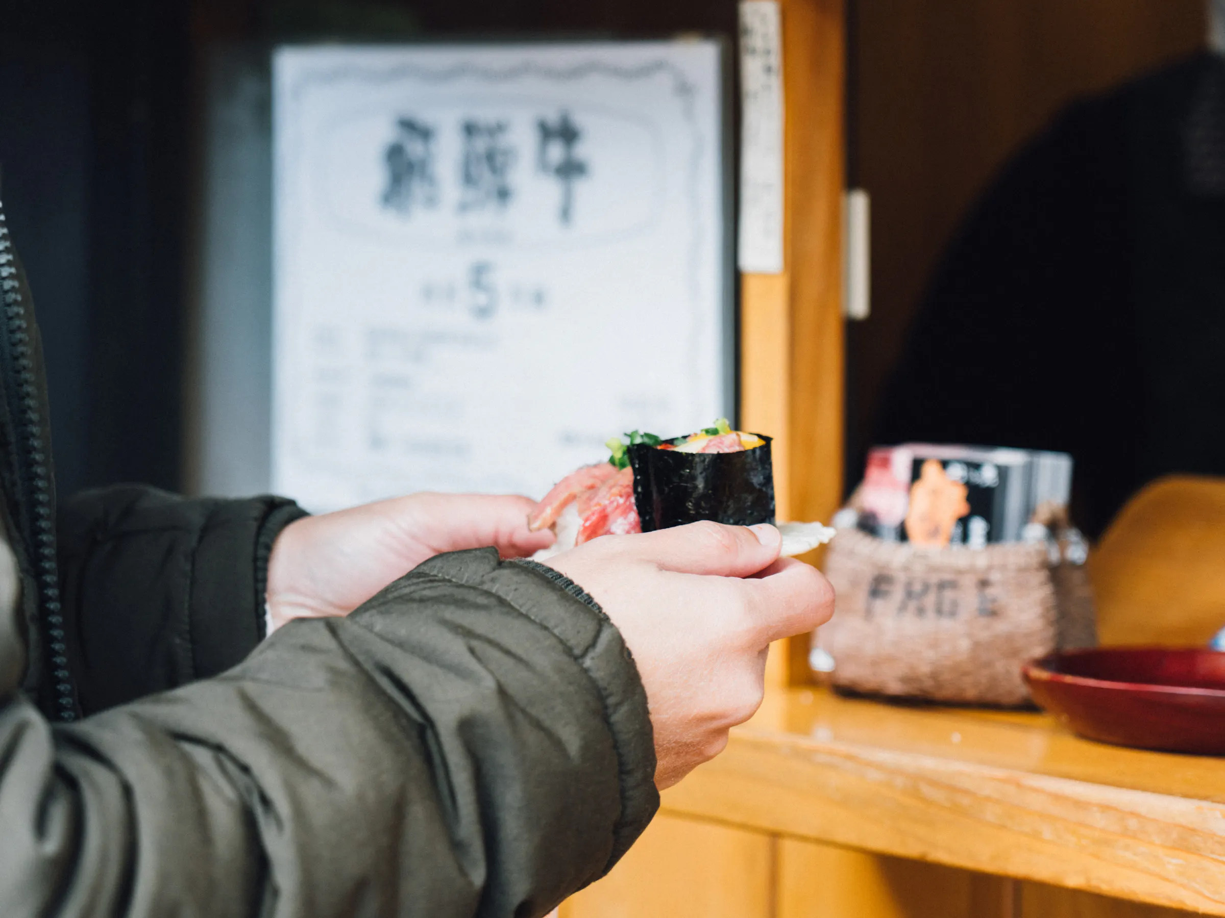 A diner holds a sushi hand roll at a wooden counter in Takayama, with small bowls, menus, and a blurred figure behind.