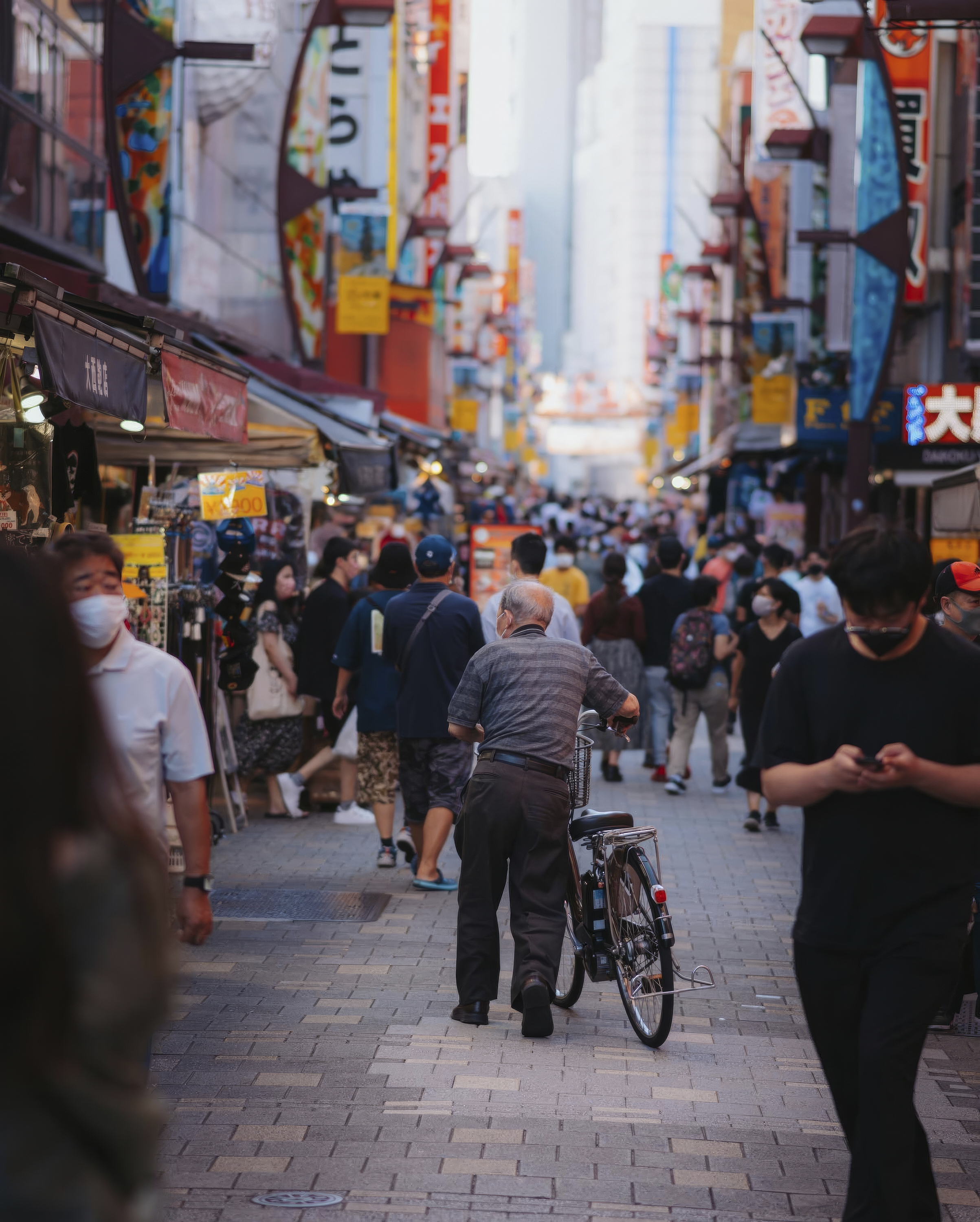 A busy Tokyo market street fills with evening pedestrians, as an older man walks a bicycle through the center of the lane.