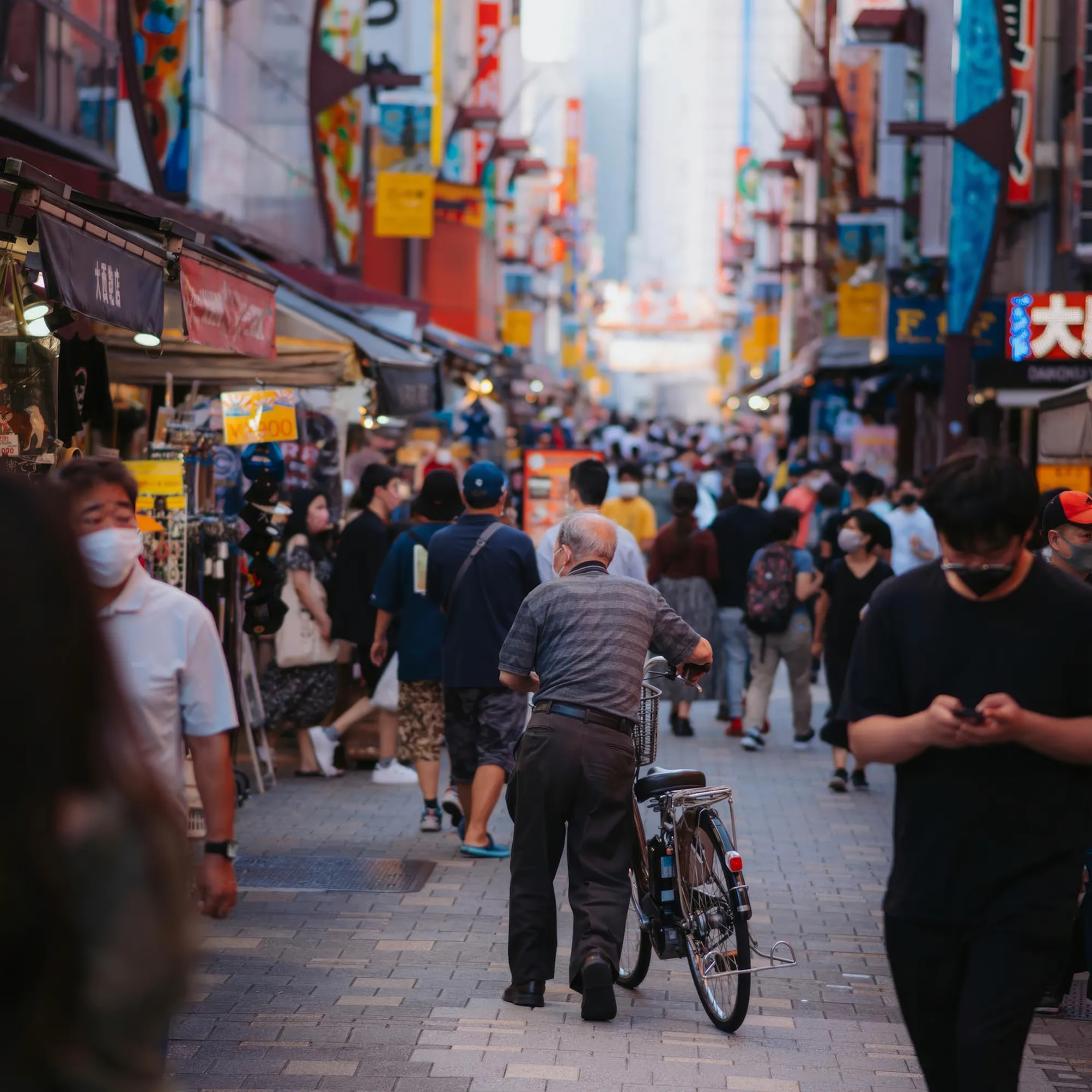 A busy Tokyo market street fills with evening pedestrians, as an older man walks a bicycle through the center of the lane.