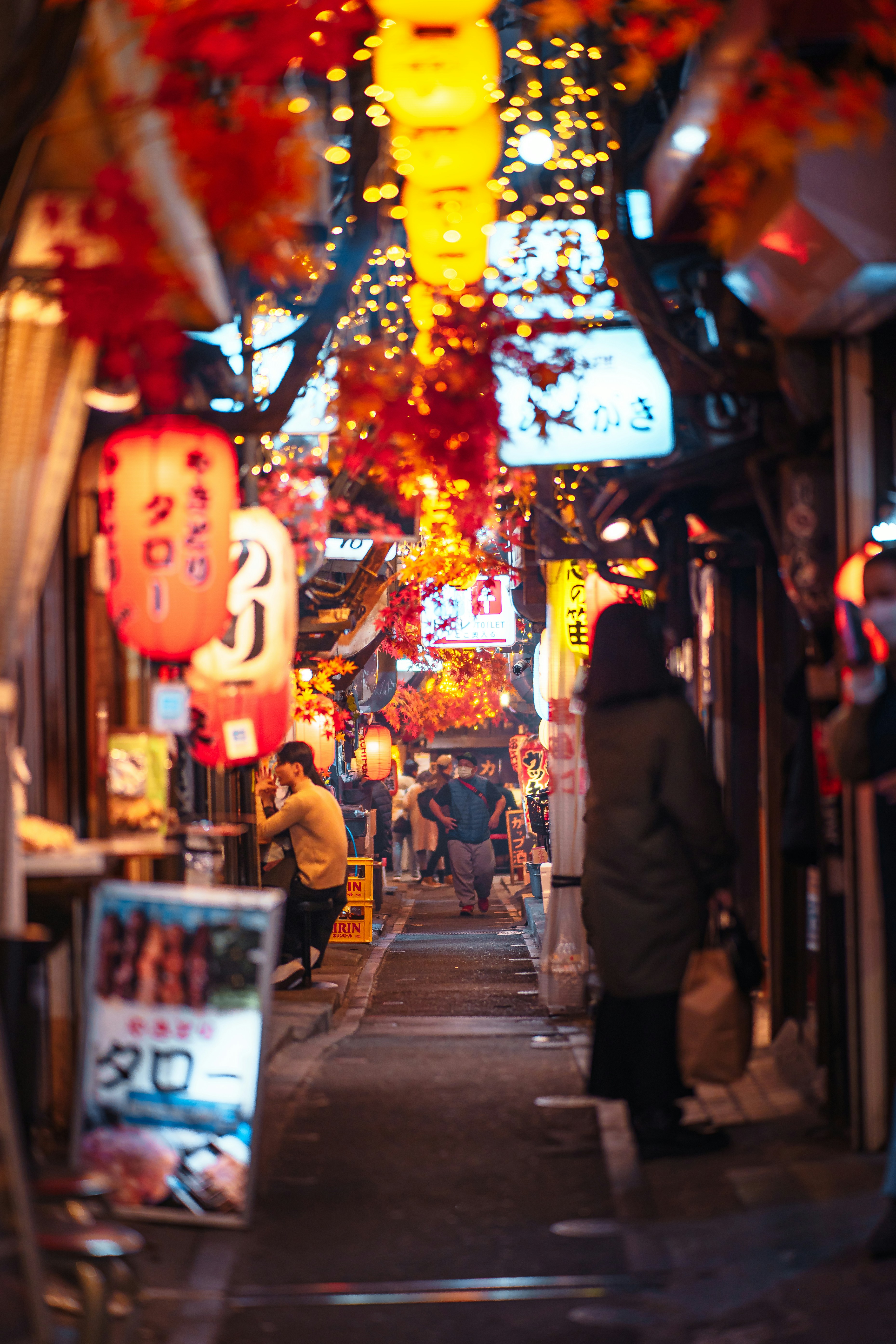 Warm lanterns and autumn leaves line a narrow alley of small bars, where diners and passersby gather in soft light.