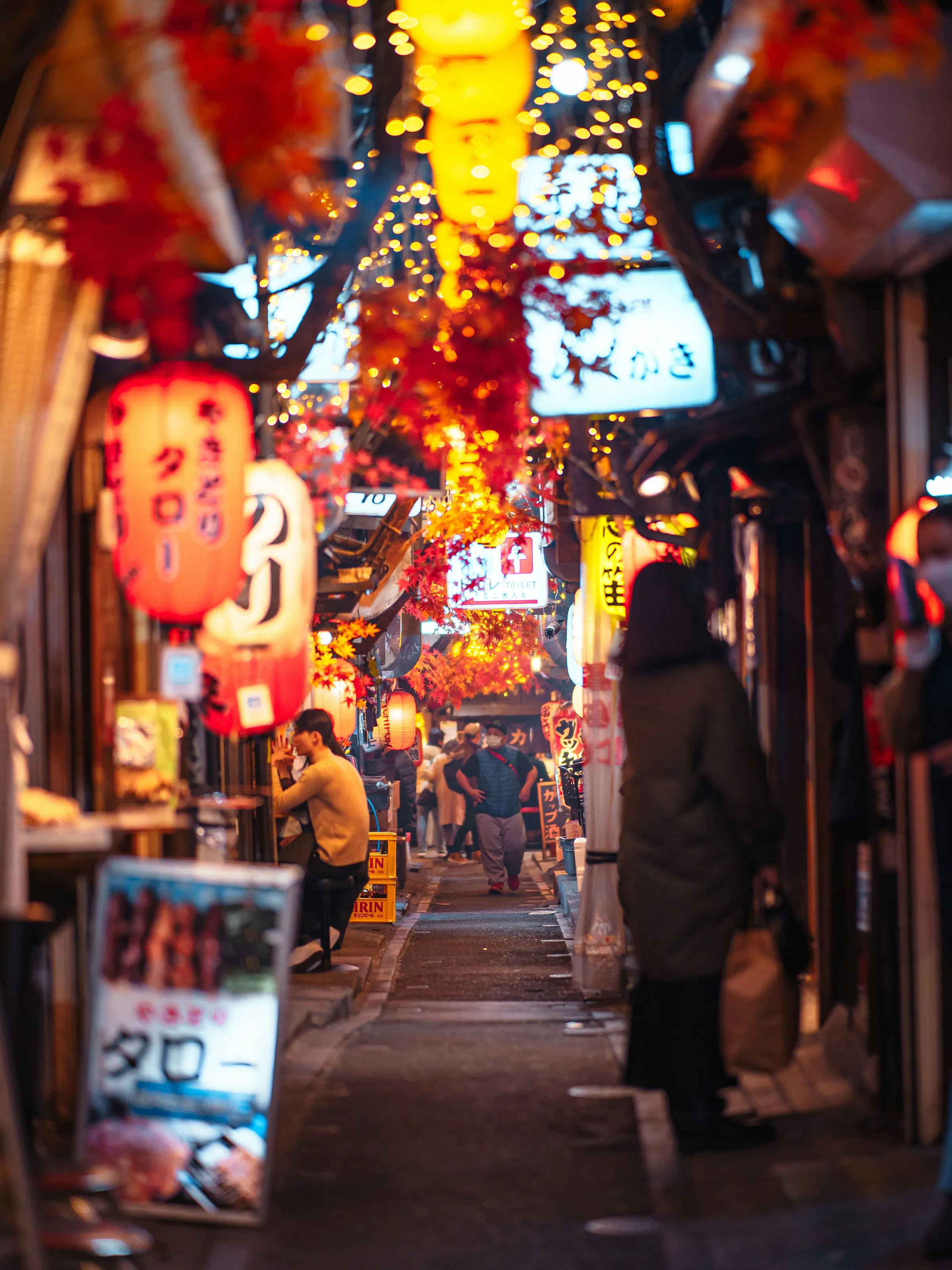 Warm lanterns and autumn leaves line a narrow alley of small bars, where diners and passersby gather in soft light.