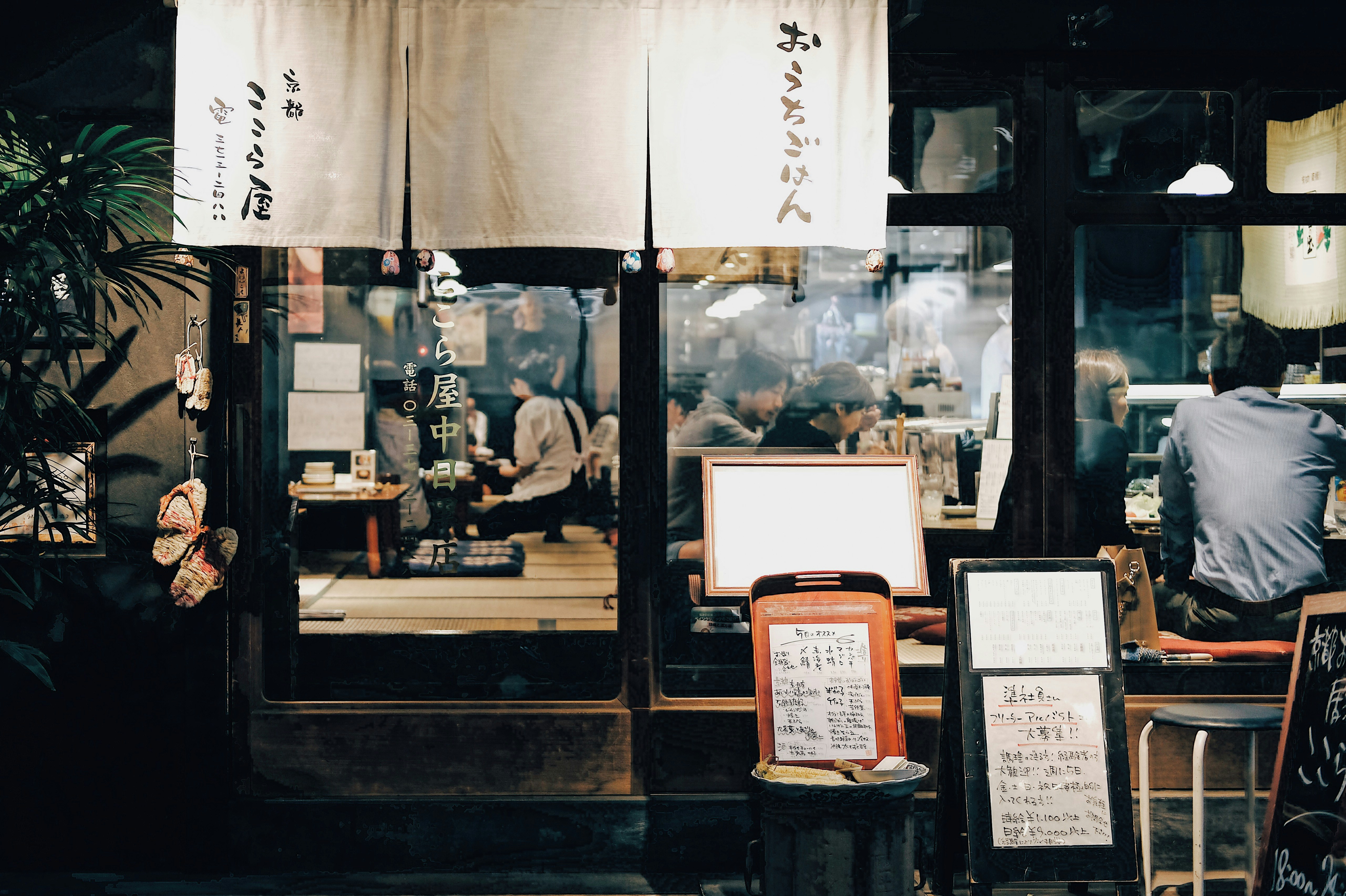 A Tokyo restaurant facade is framed by noren curtains and handwritten menus, with diners seated inside under warm light.