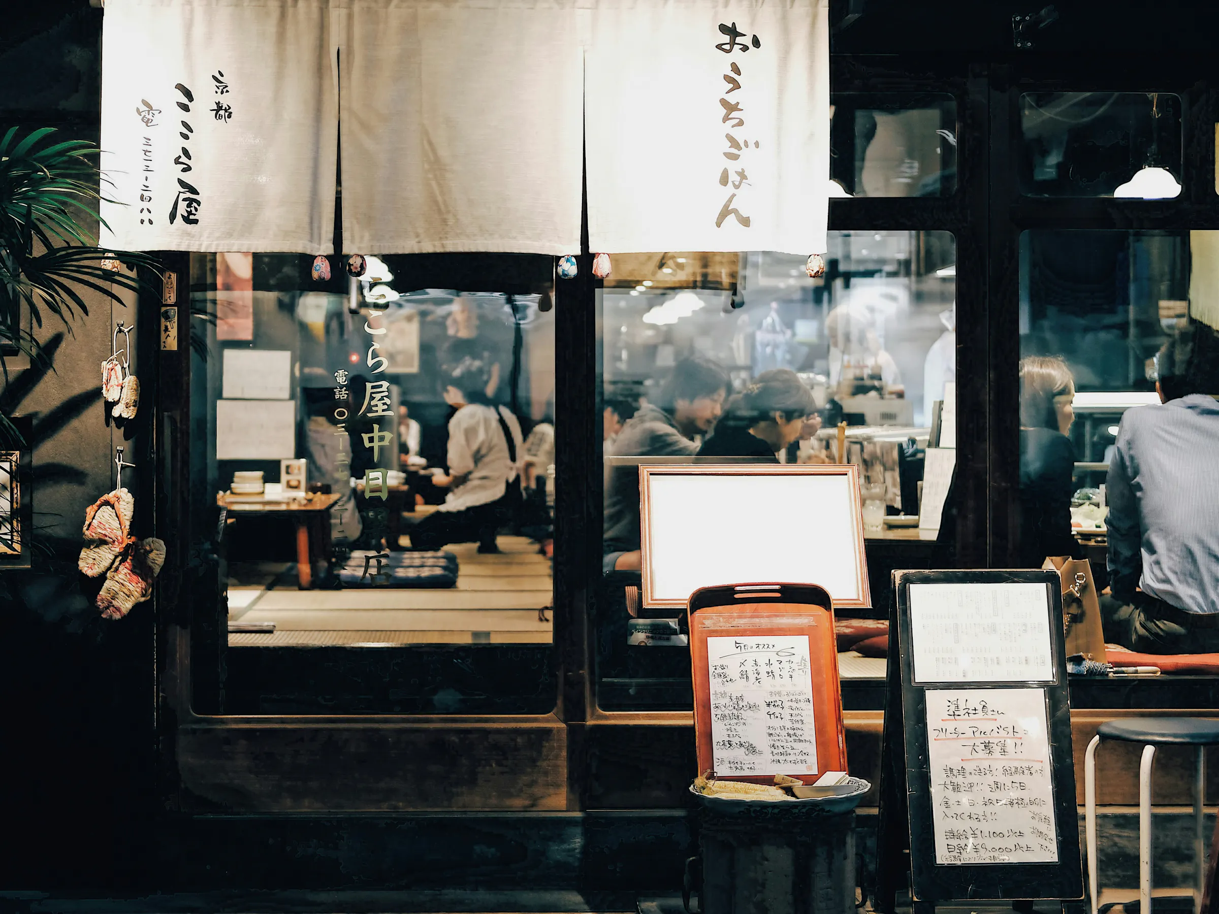 A Tokyo restaurant facade is framed by noren curtains and handwritten menus, with diners seated inside under warm light.