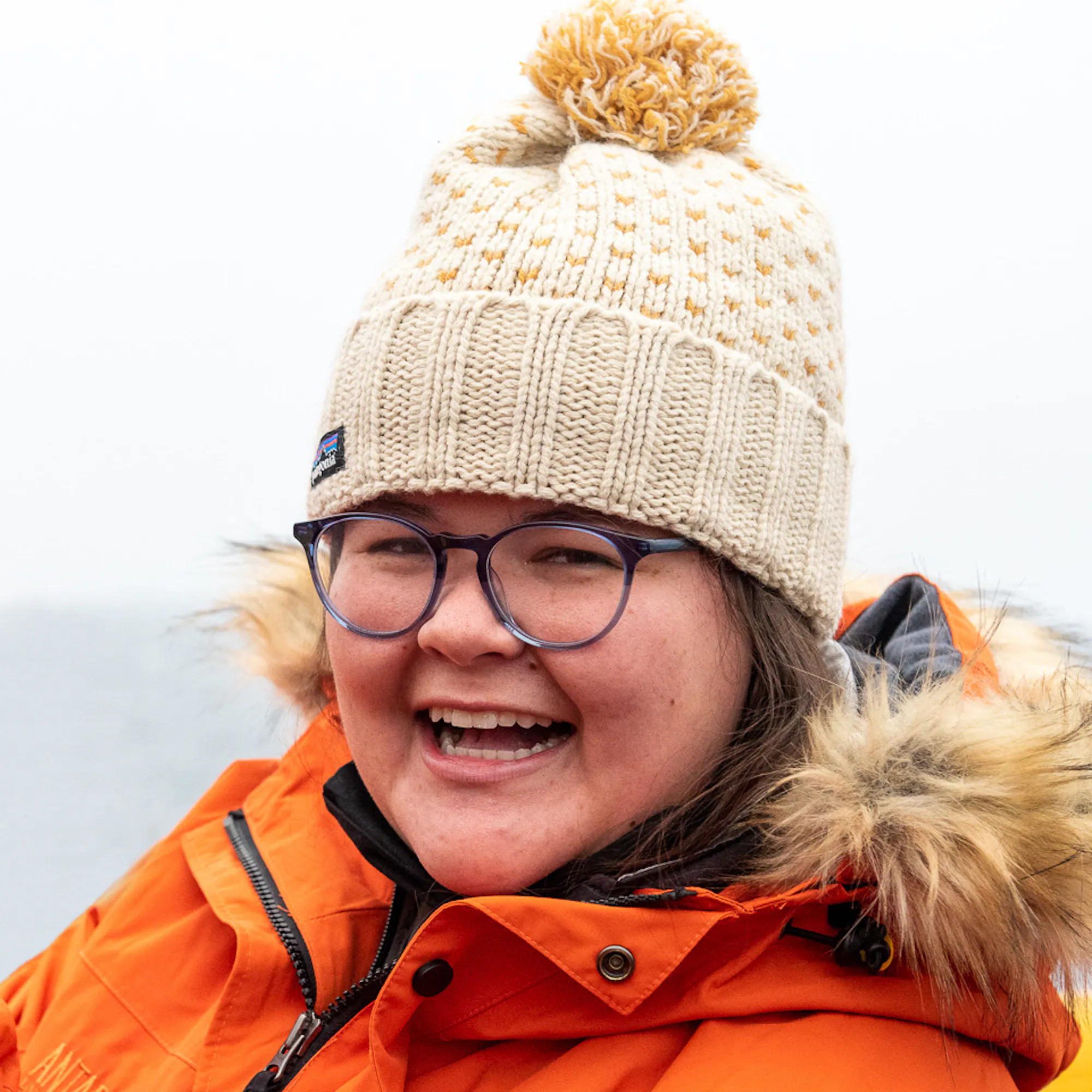 A woman wearing a white beanie and puffy orange coat smiles at the camera. 
