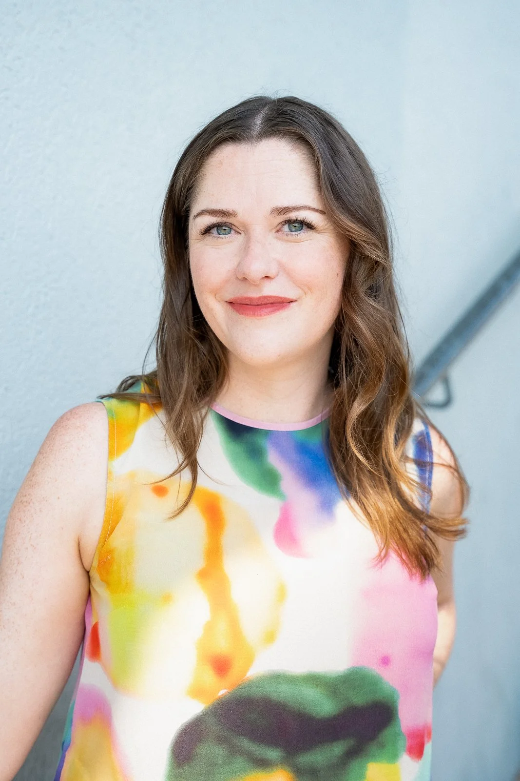 A woman with long brown hair wearing a colorful top smiles at the camera.