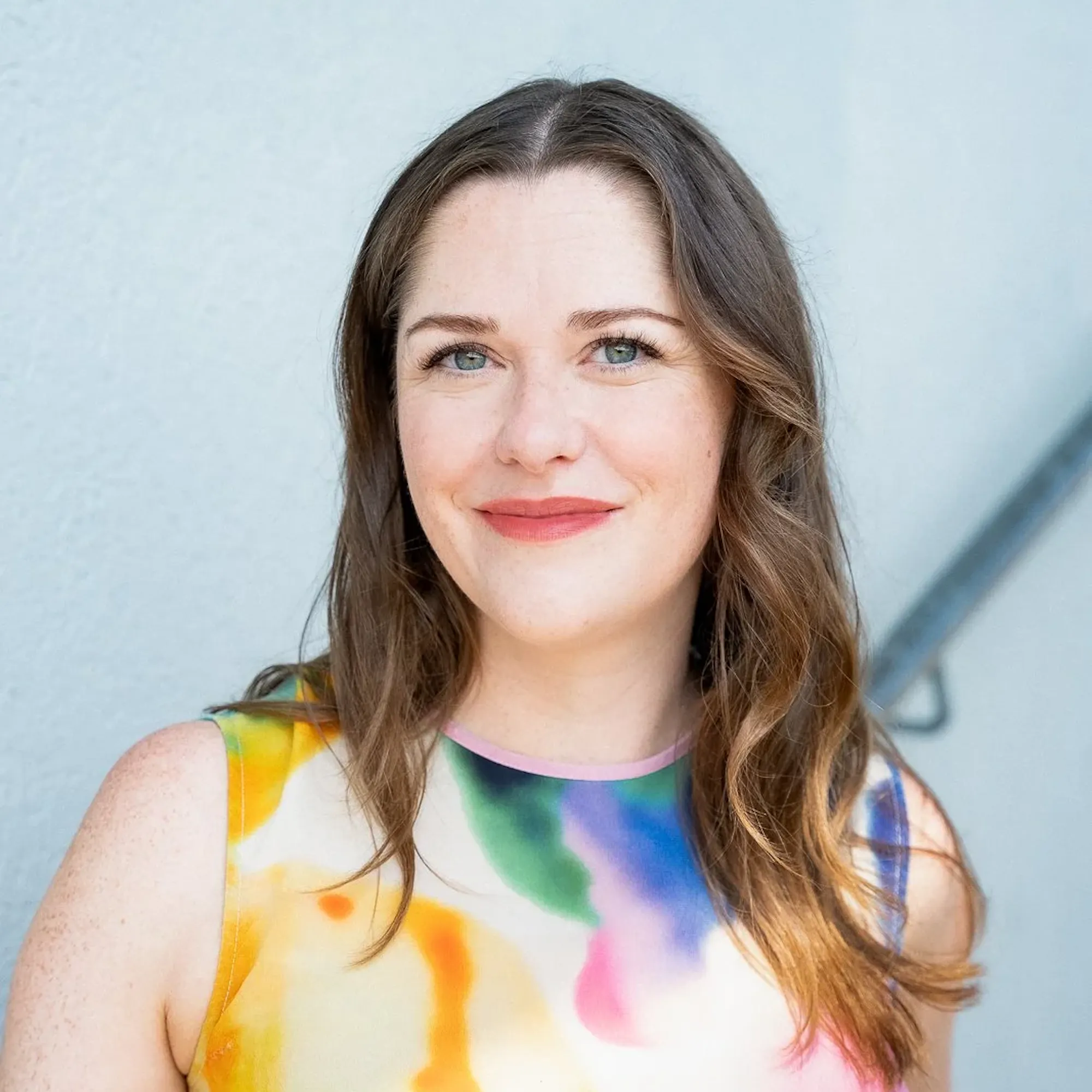 A woman with long brown hair wearing a colorful top smiles at the camera.