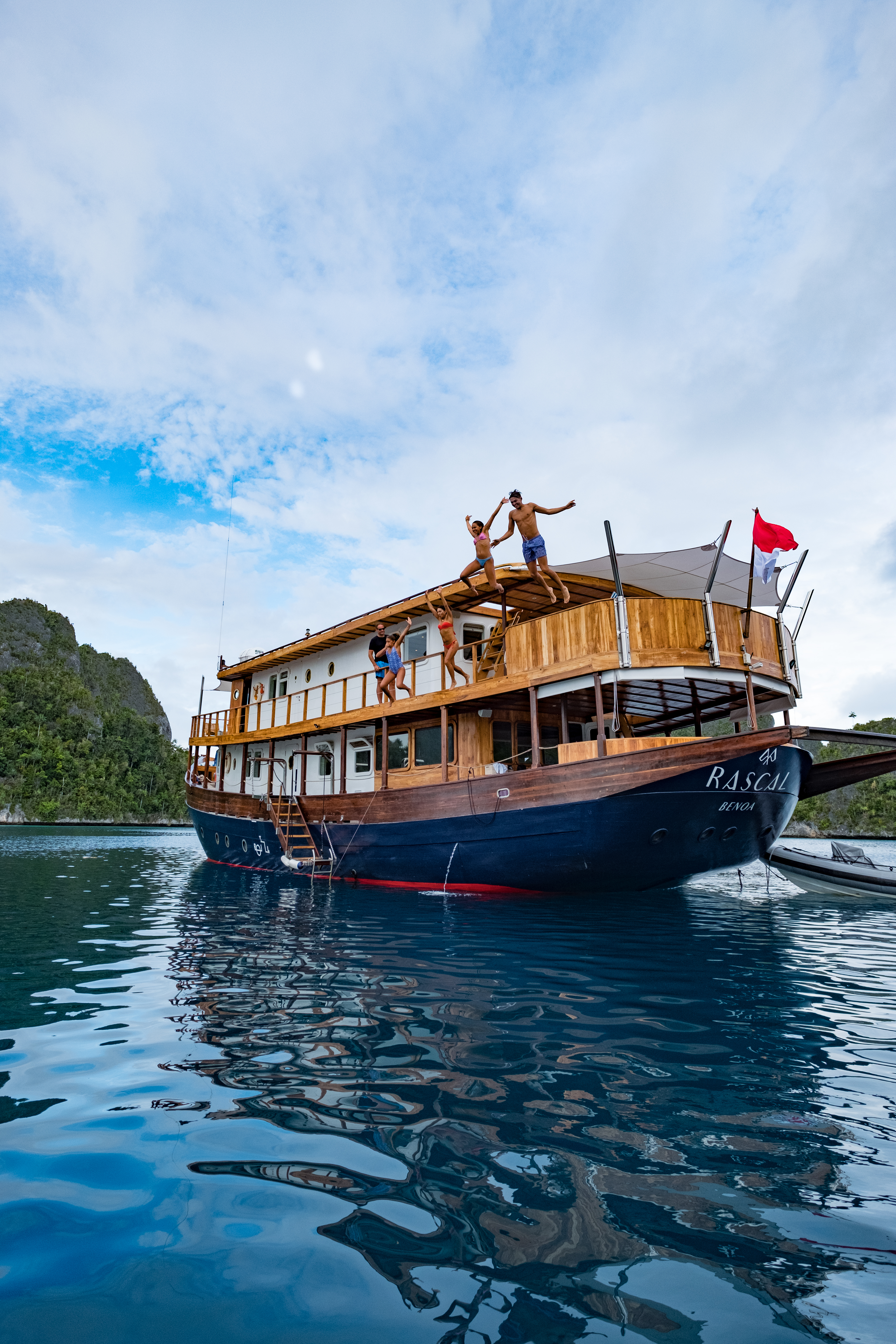 Guests leap from Rascal's upper deck into still blue water, with limestone islands rising beyond the yacht.