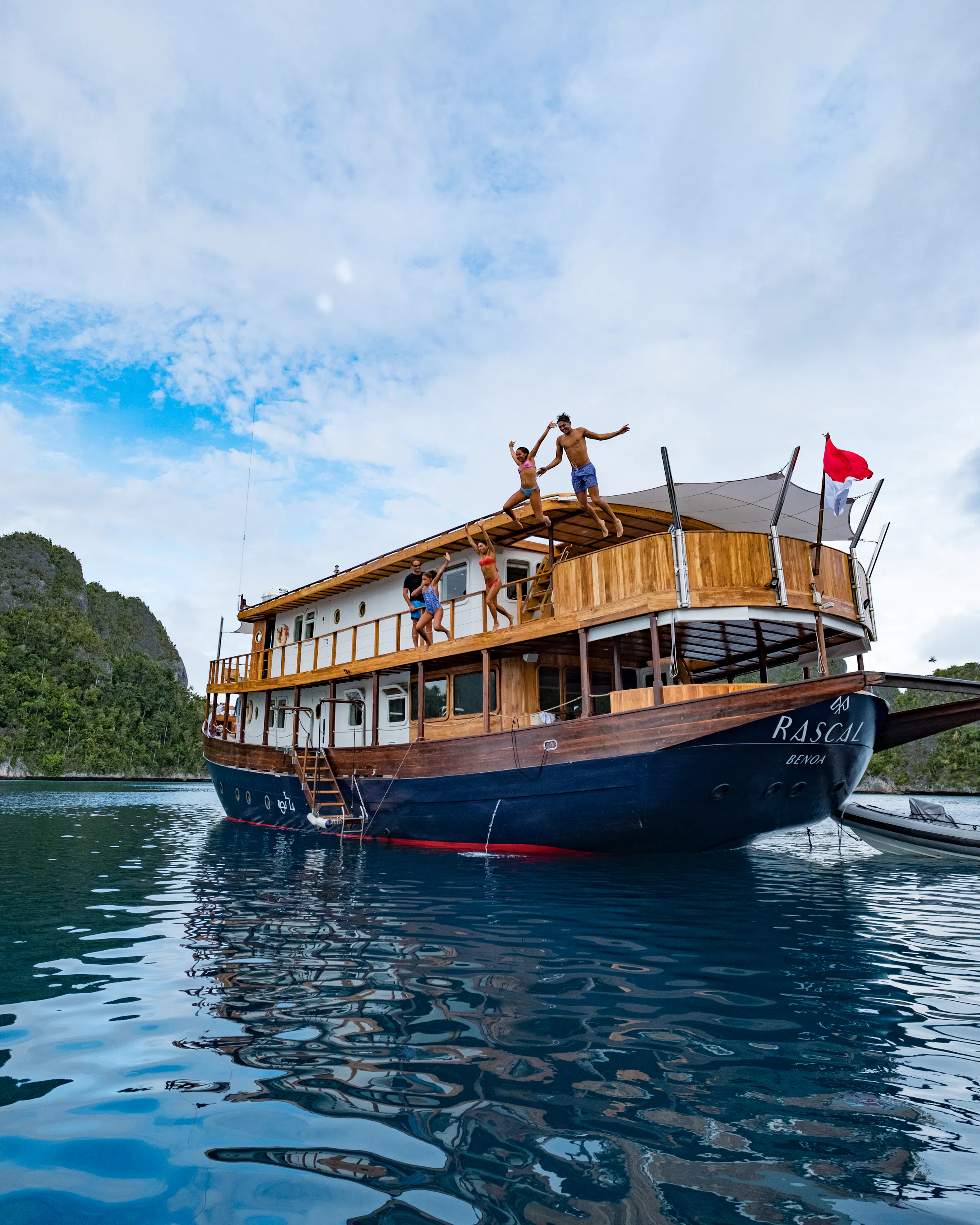Guests leap from Rascal's upper deck into still blue water, with limestone islands rising beyond the yacht.