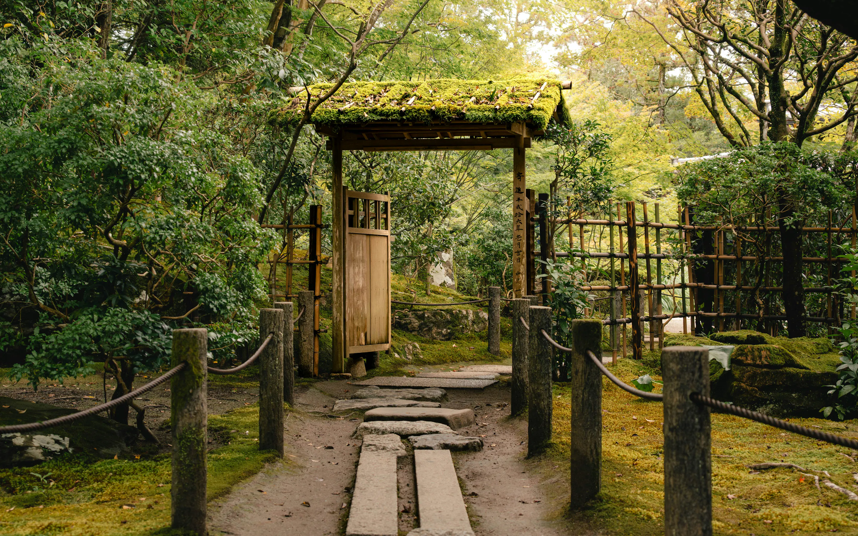 Stone steps lead to a small wooden gate framed by moss, trees, and rope-lined paths in a quiet Kyoto garden.