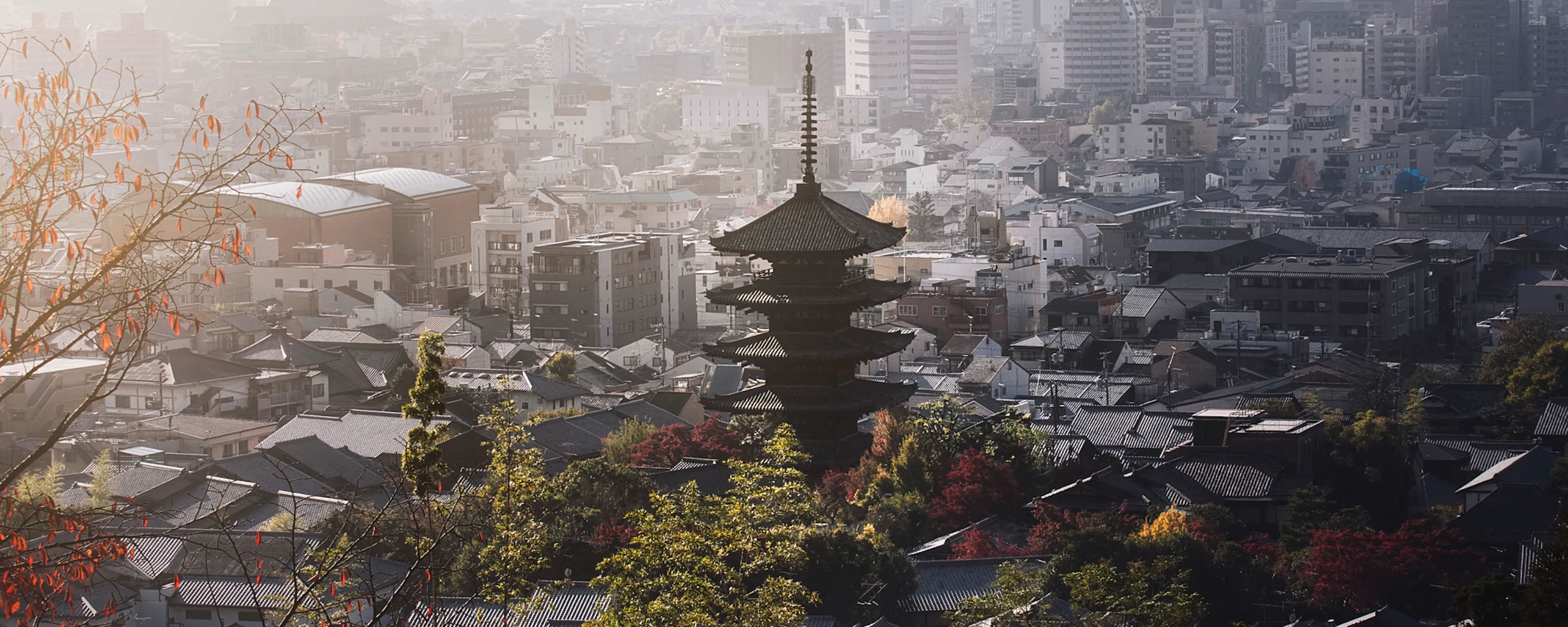 A five-story pagoda rises above Kyoto rooftops, framed by autumn foliage and a hazy mountain skyline in soft light.