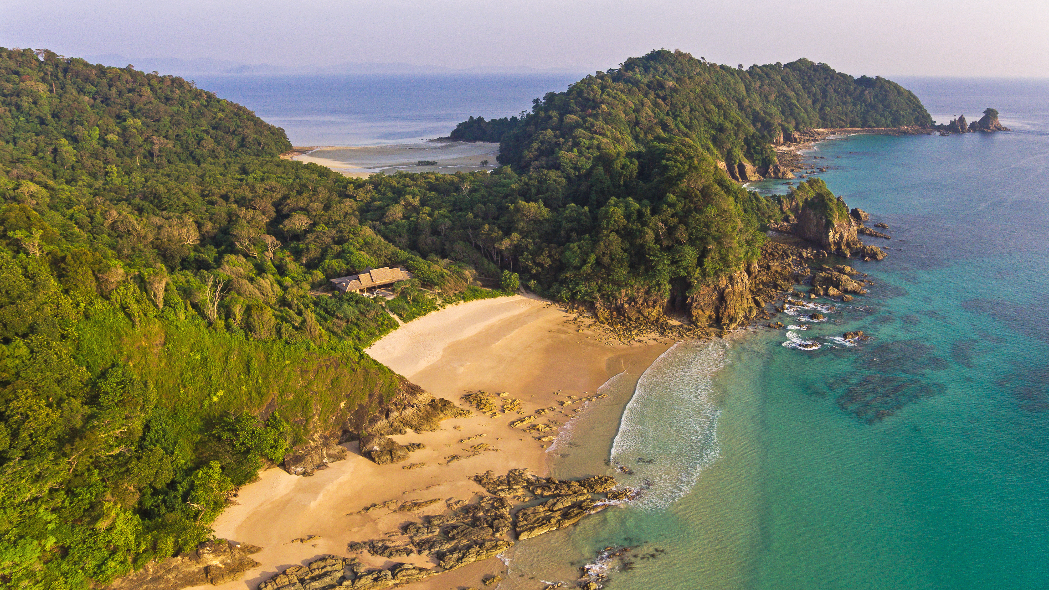 An aerial view shows a secluded beach and pavilion below steep forested headlands beside clear turquoise water.