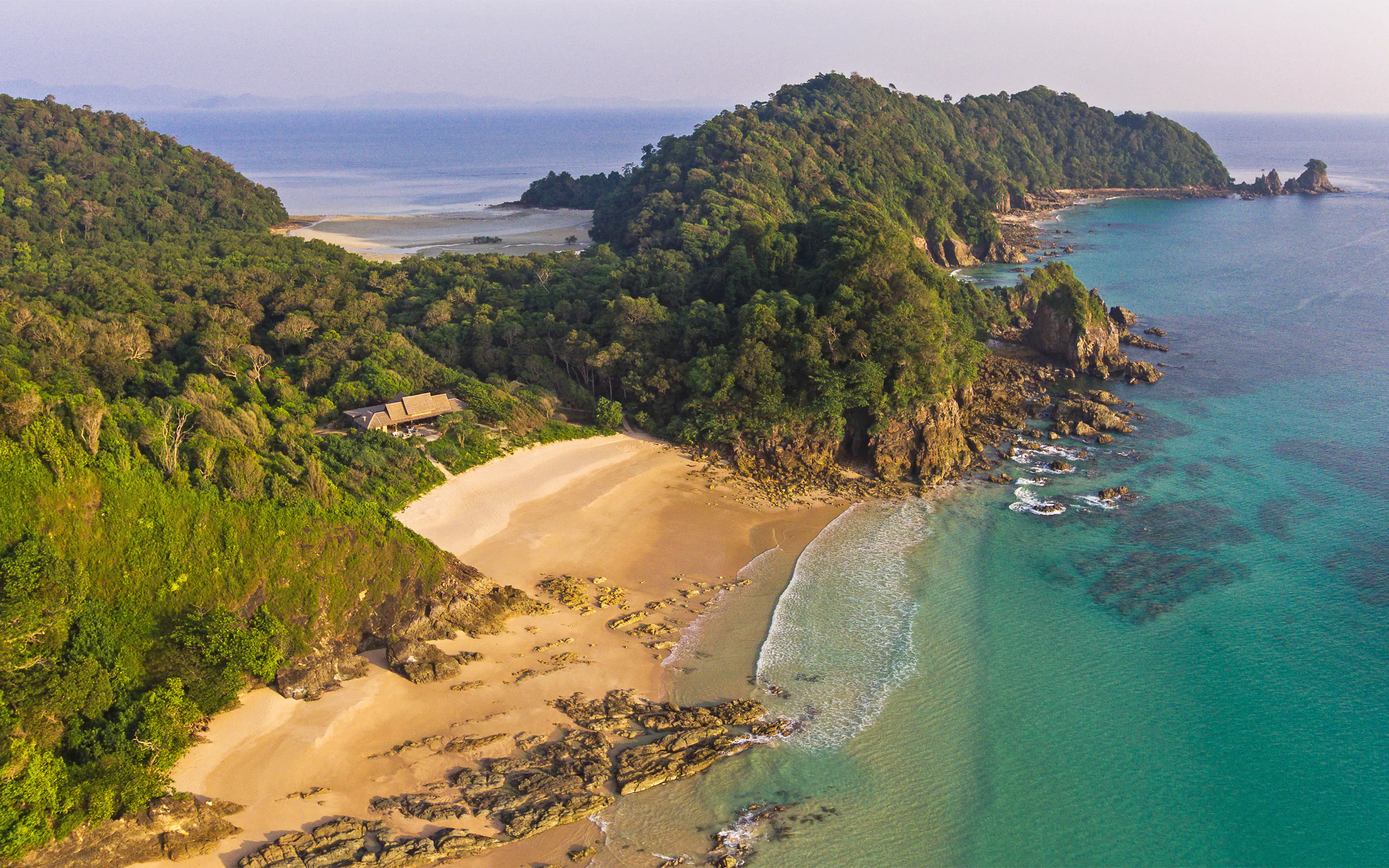 An aerial view shows a secluded beach and pavilion below steep forested headlands beside clear turquoise water.